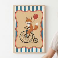A light wood-framed vertical illustration of a whimsical cartoon fox riding a vintage-style high-wheel bicycle. The fox wears a small striped bowtie and holds a single red balloon. This central scene is enclosed within a decorative red and cream scalloped border. The overall background consists of vertical blue and cream stripes. The frame is mounted on a plain white wall, with a person's shoulder and arm partially visible in the lower right corner.
