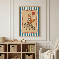 A framed illustration of a fox riding a vintage bicycle hangs on a neutral wall. The fox wears a small blue bow tie and holds a red balloon. The artwork features a scalloped beige center set against blue and white vertical stripes. Below the frame, a wooden shelf contains woven baskets, colorful books, and a wooden toy train. A portion of a light-colored armchair with a fuzzy blanket is visible on the right side.