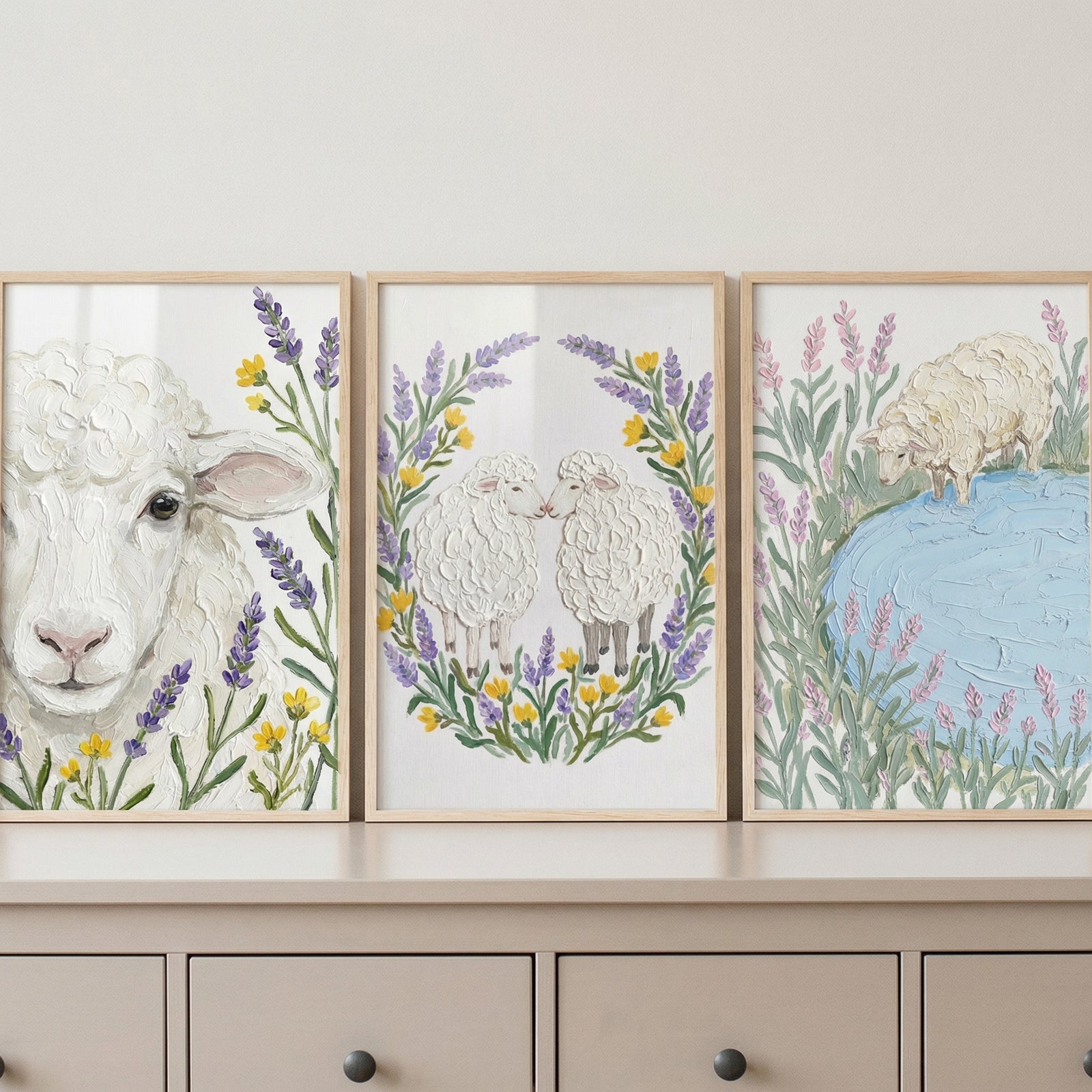 Three framed artworks featuring white sheep are displayed side-by-side on a neutral-toned dresser. Each piece has a heavy, textured finish that gives the wool and petals a three-dimensional look. The left art shows a close-up sheep portrait among lavender and yellow flowers. The middle art depicts two sheep in a floral wreath. The right art features a sheep by a blue pond surrounded by tall purple stalks. The light wood frames complement the soft, pastel color palette of the series.