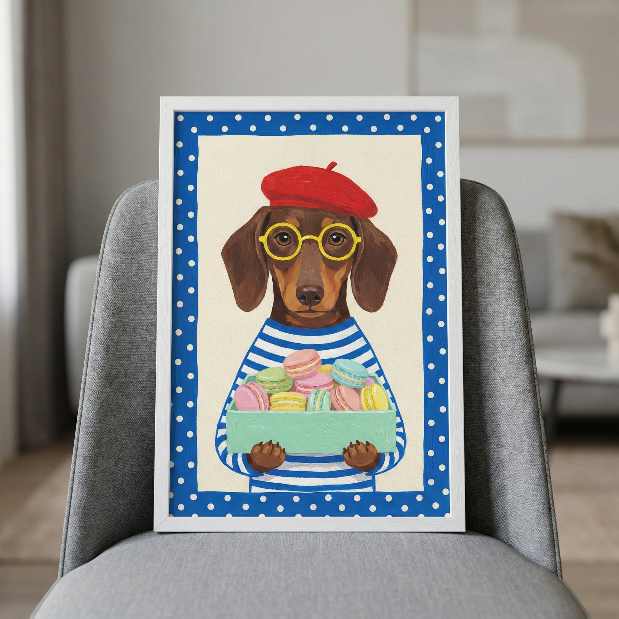A framed illustration of a brown dachshund wearing a red beret, yellow round-rimmed glasses, and a blue and white striped shirt. The dog is holding a light green box filled with various pastel-colored macarons. The artwork is bordered by a blue frame with white polka dots and is displayed in a thin white frame leaning against a gray fabric chair in a modern room.