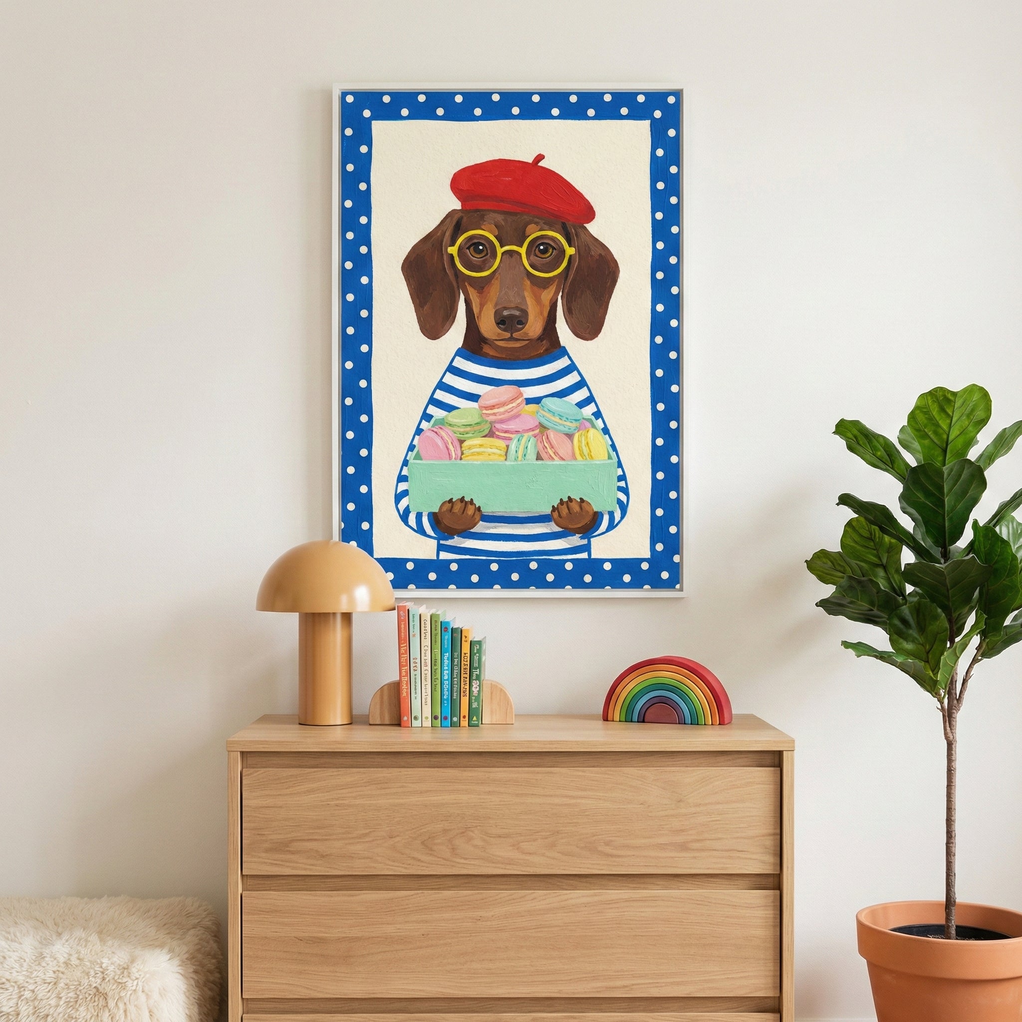 A framed vertical illustration of a brown dachshund dog hangs on a white wall above a light wood dresser. The dog is depicted wearing a red beret, yellow-rimmed glasses, and a blue and white horizontally striped shirt. It holds a small light green box filled with colorful, round, multi-layered treats. The artwork has a wide blue border with white polka dots. On the wooden dresser below the frame are a tan mushroom-style lamp, several small colorful books, and a wooden rainbow arch toy. A tall green potted p