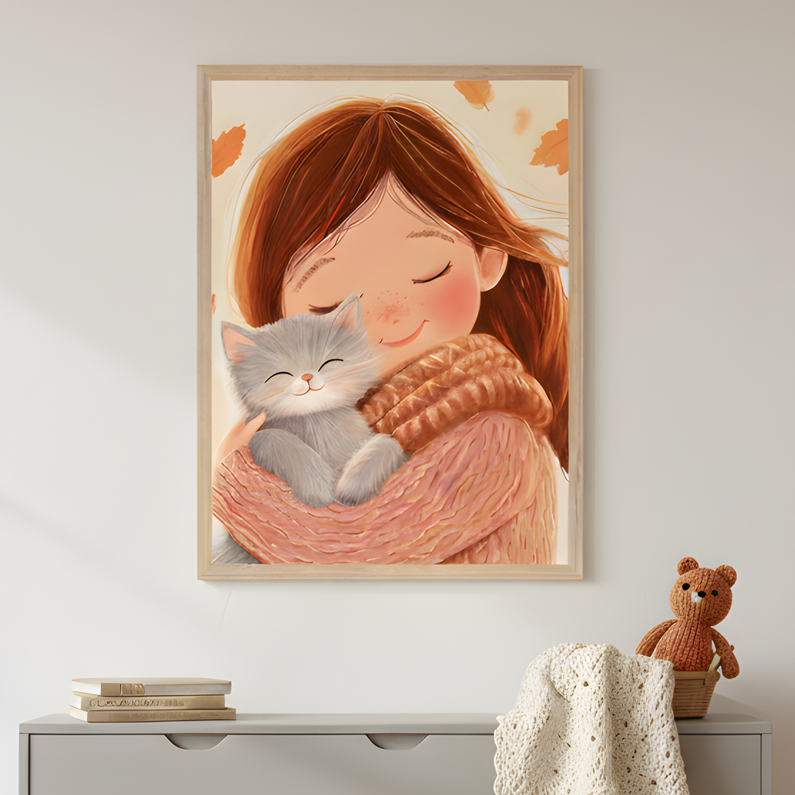 Framed illustration of a smiling girl with long brown hair hugging a fluffy gray kitten, both with closed eyes, wrapped in cozy knit sweaters. The warm autumn-themed artwork hangs above a dresser with stacked books, a knitted teddy bear, and a cream-colored blanket, adding a soft and comforting feel to the space.