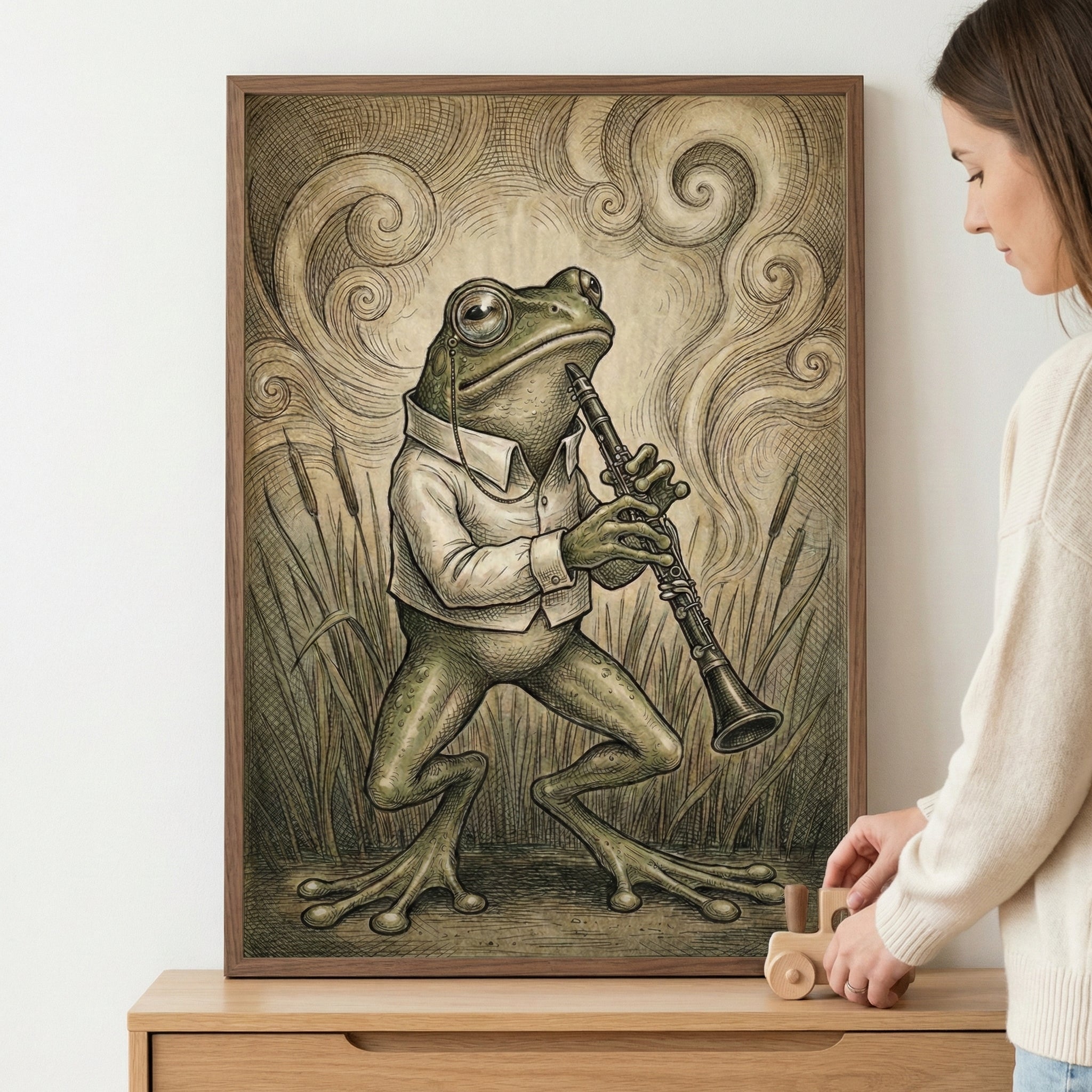 Framed illustration of a frog wearing a button-up shirt and monocle, playing a clarinet amid tall grass with swirling background lines adding a dreamy, vintage touch. A person is seen placing the artwork on a wooden dresser, holding a small wooden duck toy, suggesting a cozy and curated space.
