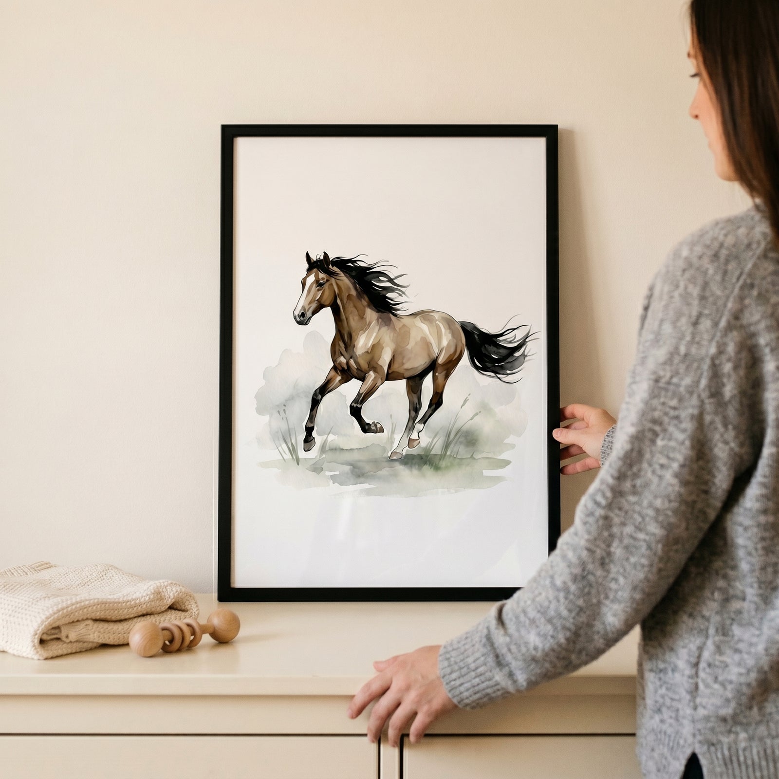 A person holds a framed galloping brown bay horse wall art piece above a dresser with a wooden baby rattle.