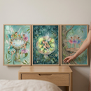 Three framed, whimsical illustrations are arranged side-by-side on a light wood nightstand against a neutral-colored wall. Each artwork features a different scene of delicate, winged sprites in an ethereal forest setting.

The illustration on the left shows several winged figures gathered around a stone fountain, some perched on the rim and others hovering nearby. 