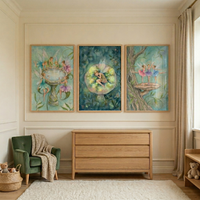 A serene nursery features three framed, watercolor-style illustrations depicting whimsical woodland fairies hung above a light-wood dresser with three drawers. 

On the left, one piece of art shows several fairies with vibrant wings gathered around a stone birdbath. The center piece portrays a single fairy resting within a glowing, translucent sphere in a dark, enchanted forest. 