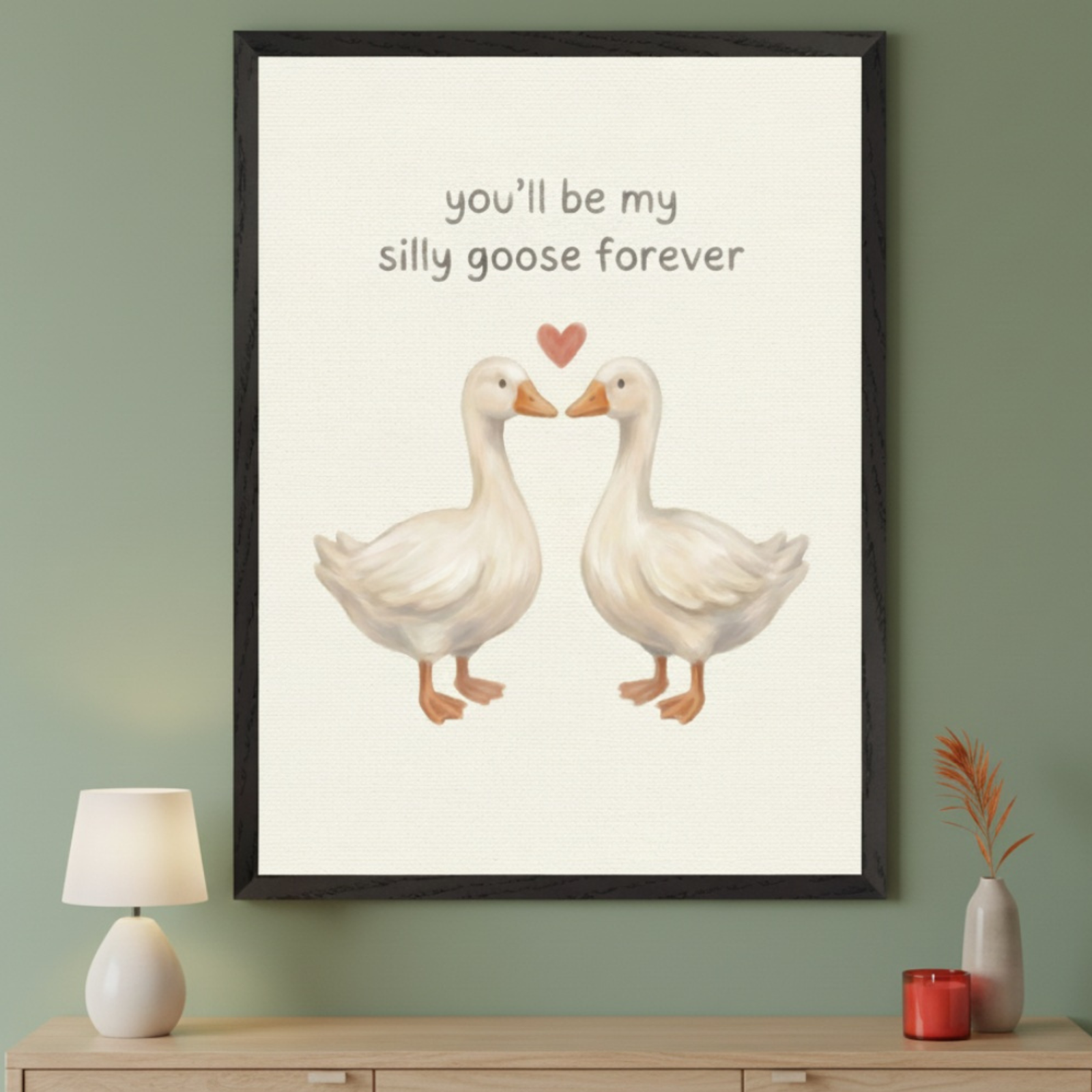 Modern room with a light wood dresser. A framed print of two white geese, a red heart, and text "you'll be my silly goose forever" in an illustration style hangs centered above the dresser.