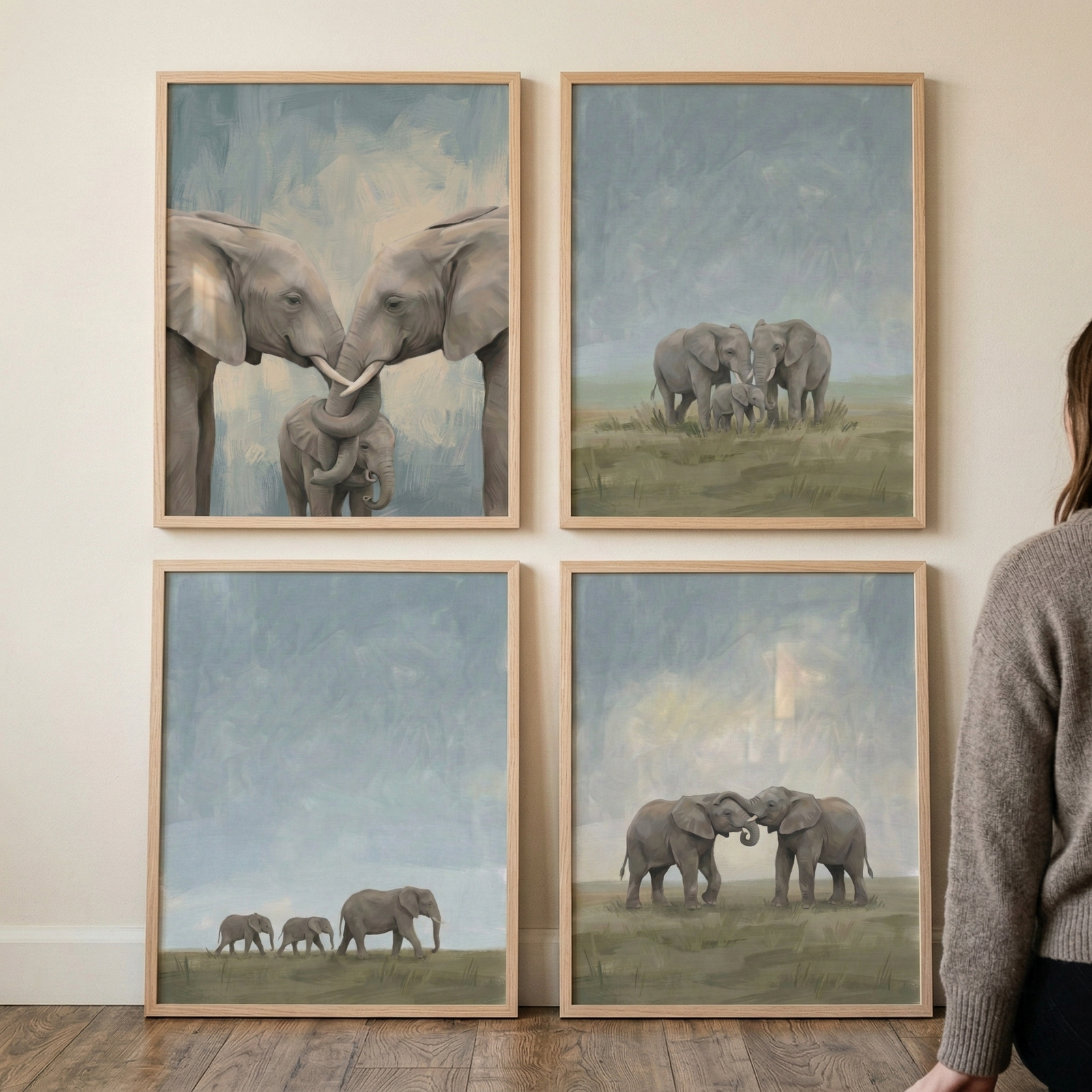 Four light-wood framed artworks are displayed in a two-by-two grid on a neutral wall. Each piece features a textured, soft-toned illustration of elephants. The top-left shows two adults with entwined trunks over a calf. The top-right depicts three elephants in a grassy field. The bottom-left shows a trio of elephants walking in a line. The bottom-right features two elephants facing each other. The color palette is a serene mix of dusty blue, grey, and muted green. A person is partially visible on the right.