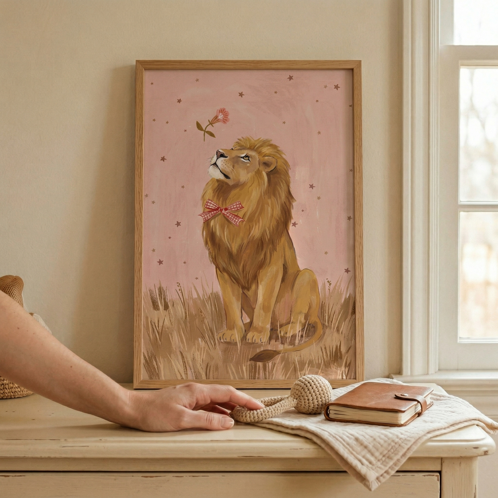 A vertical wooden frame displays a whimsical illustration of a lion with a shaggy mane sitting in tall grass. The lion wears a pink checkered bow and gazes at a pink flower in a star-filled rose sky. The frame sits on a light wooden dresser next to a folded cloth holding a brown leather journal and a knitted rattle. A person's hand reaches toward these items. A beige wall and white window frame create a soft, warm background.