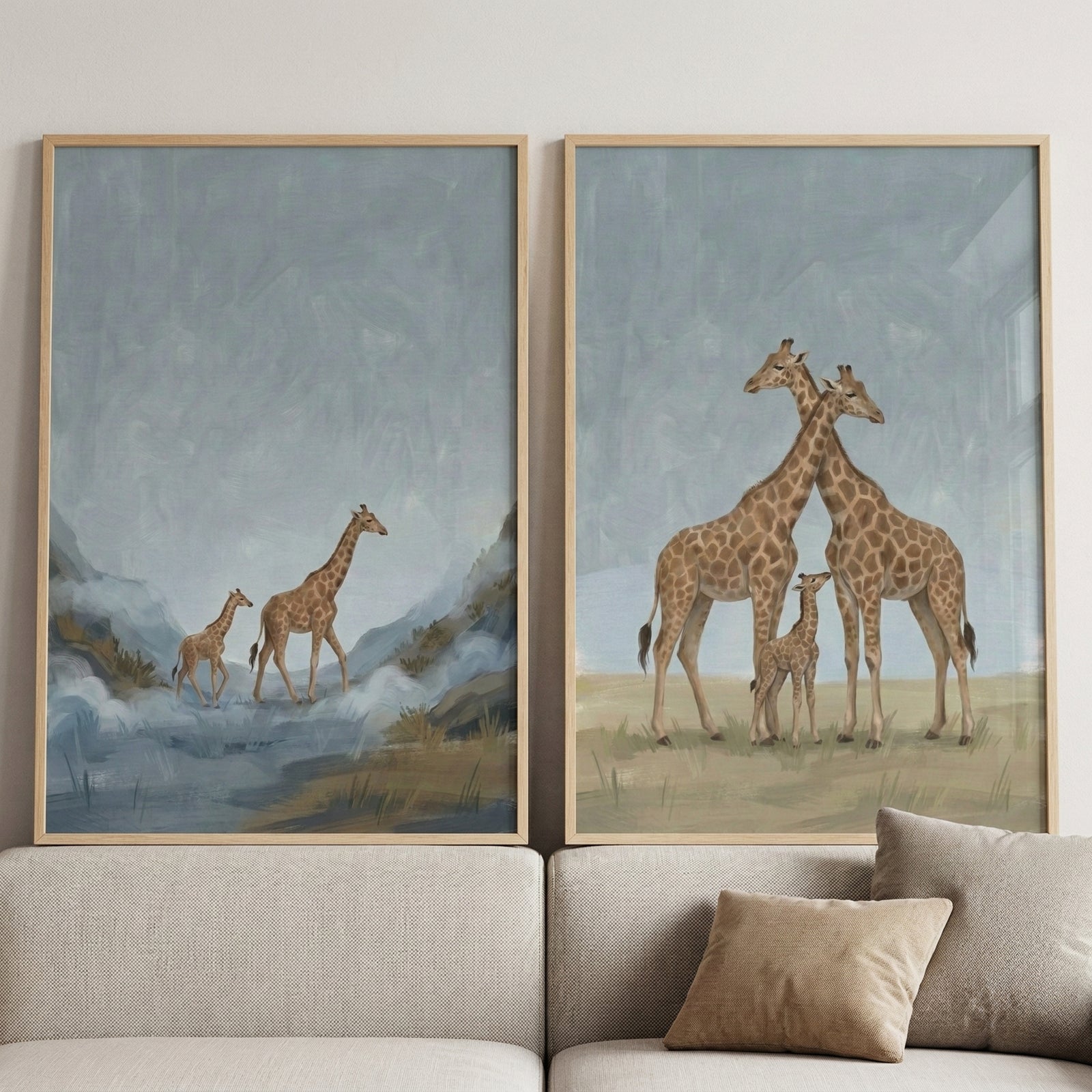 Two large framed giraffe paintings lean against a light colored sofa with neutral beige pillows. One painting shows an adult and baby giraffe walking through a misty blue landscape, while the other shows two adult giraffes with a calf standing together on pale grass.