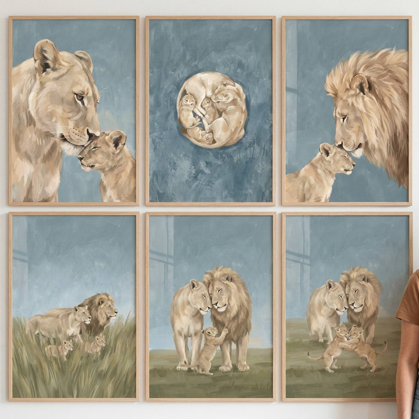 Six framed wall decorations are arranged in a three-by-two grid. Each piece features lions in a soft, illustrative style with a palette of muted blues, warm tans, and earthy greens. The top row shows a lioness nuzzling a cub, a group of cubs huddled in a circle, and a male lion touching noses with a cub. The bottom row depicts a pride in tall grass, a cub reaching up to its parents, and two cubs playing while two adults watch. The frames are made of light-toned wood.