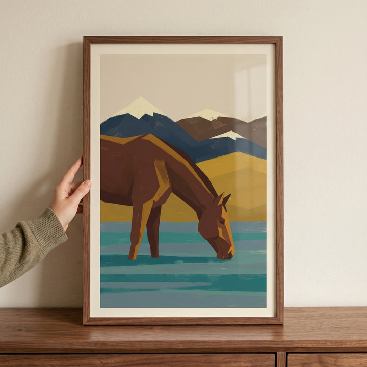 Hand holds framed geometric teal water horse wall art of a horse drinking against a mountain landscape.