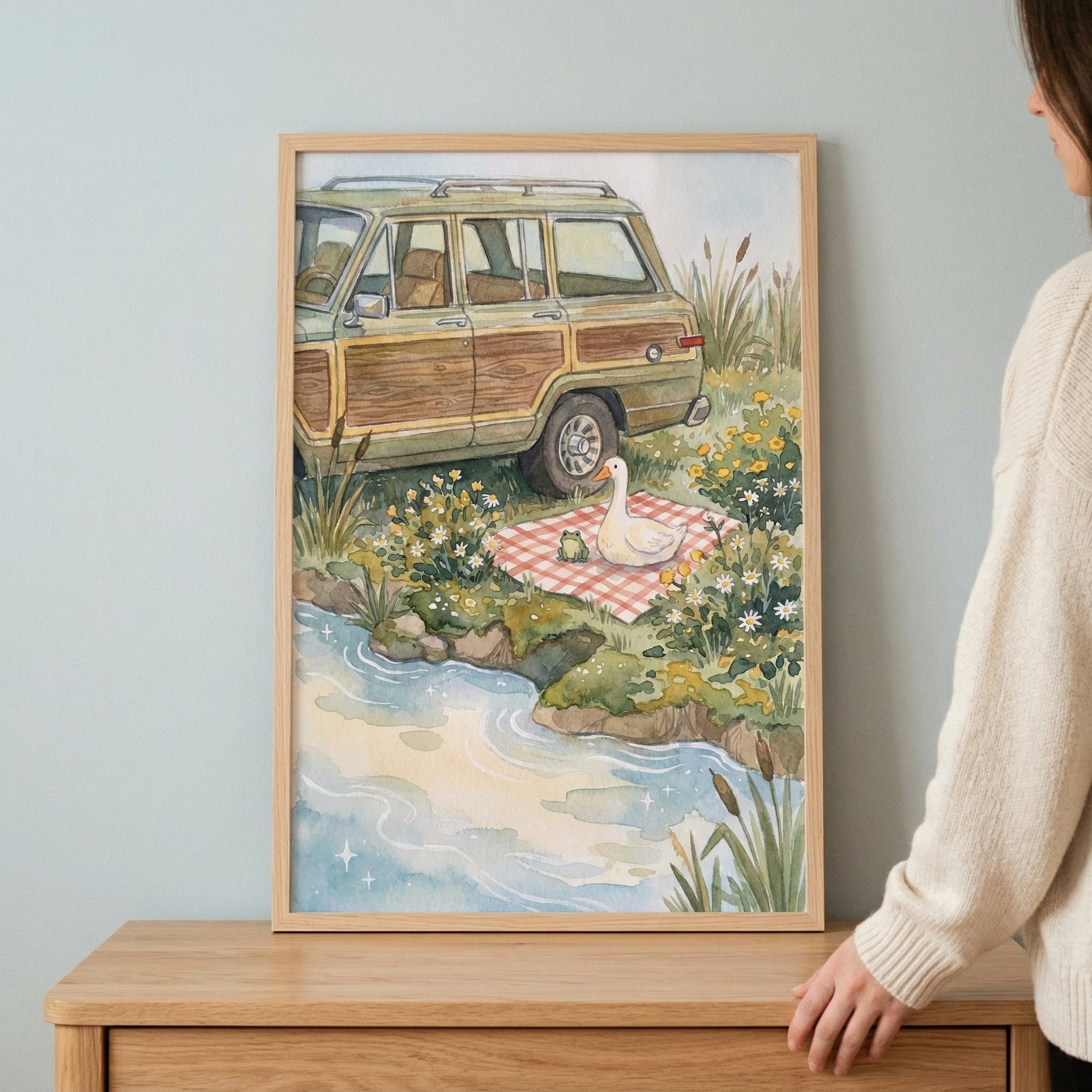 An illustration in a light wood frame is held by a person wearing a cream-colored knit sweater. The artwork depicts a vintage station wagon with wood-grain side panels parked in a field of tall grass and yellow wildflowers. 