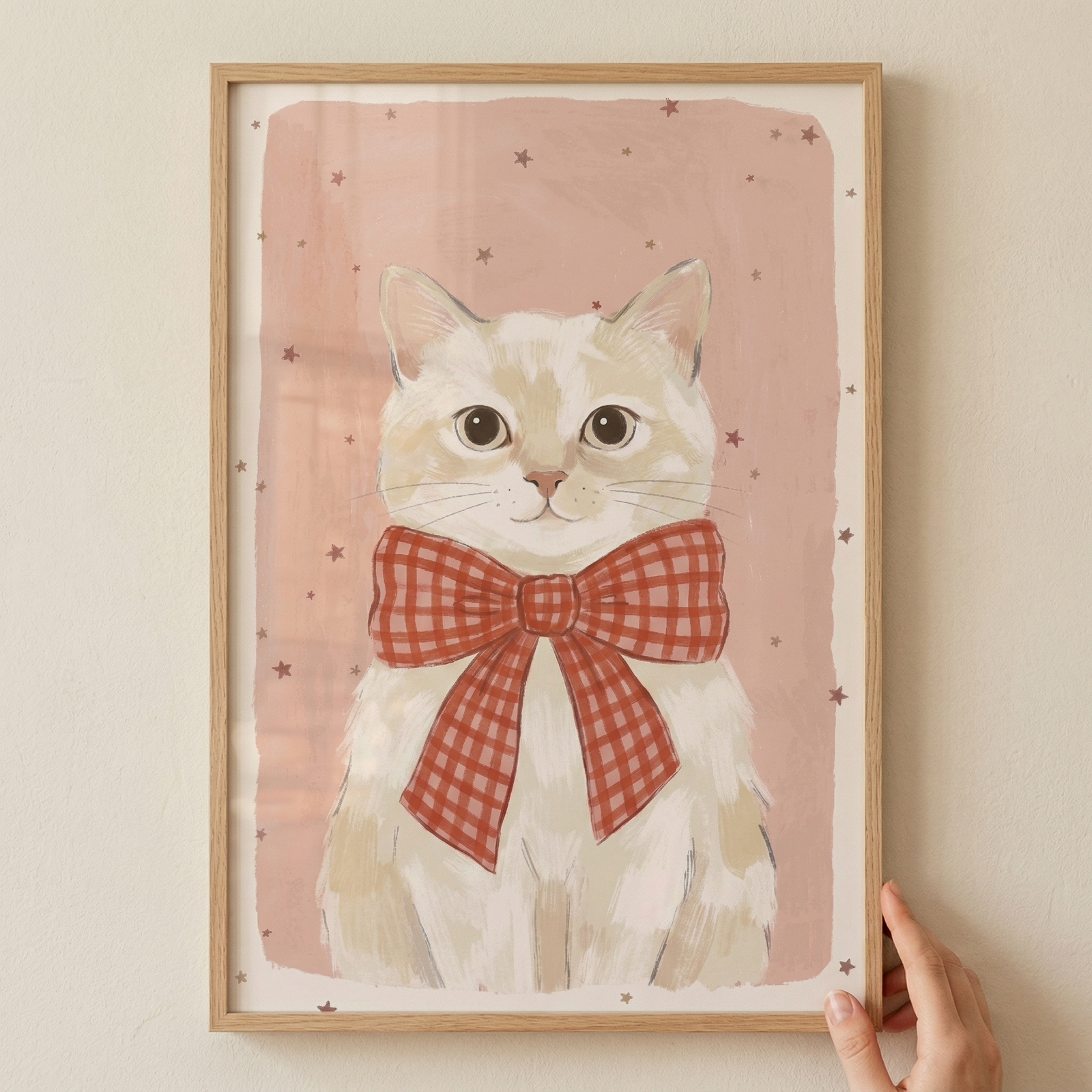 A framed illustration features a light-colored cat with large, dark eyes and a prominent red and white checkered bow tie. The subject is centered against a soft, muted pink backdrop decorated with tiny, scattered star shapes. The entire piece is enclosed in a simple, light wooden frame, which is being held by a hand at the bottom right corner.