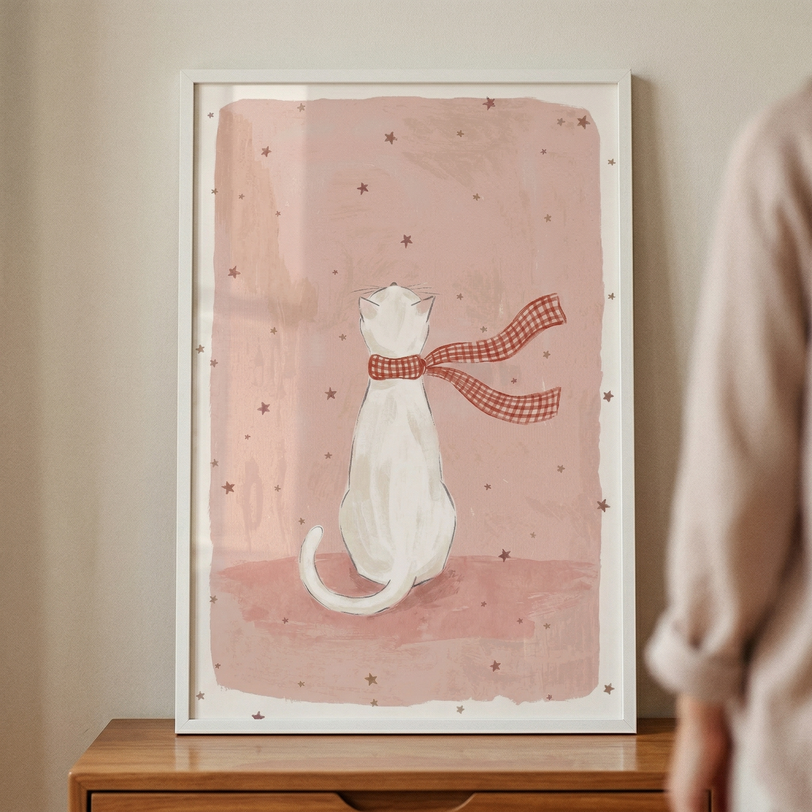 A framed illustration of a white cat seen from behind, wearing a long red and white checkered scarf that blows to the right. The cat is centered against a dusty rose, watercolor-style background dotted with small, dark stars. The artwork is encased in a simple white frame and rests on a wooden surface. A person's arm, blurred and out of focus, appears on the right edge of the image.