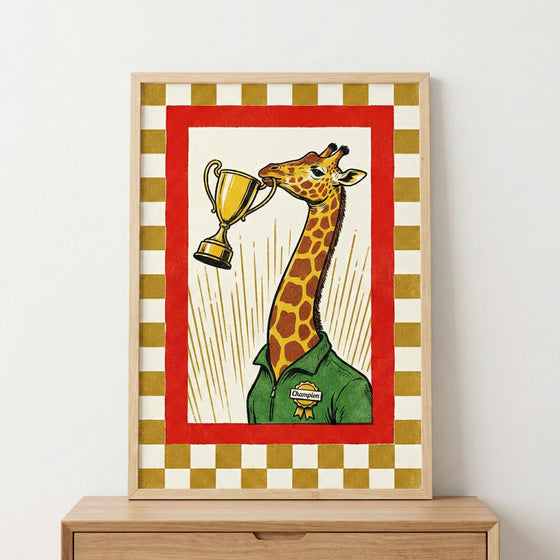 A framed graphic illustration features a giraffe wearing a green collared shirt with a yellow "Champion" badge. The giraffe holds a golden trophy cup in its mouth. Behind the giraffe are radiating yellow lines on a white background, all contained within a bold red frame. An outer border of gold and off-white checkered squares completes the design. The artwork is housed in a light wood frame, sitting atop a wooden dresser.