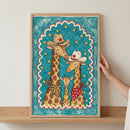 Framed illustration of a giraffe family dressed in western cowboy attire, hanging above a light wood dresser in a neutral-toned living space. The playful artwork features two adult giraffes in cowboy hats and bandanas with a baby giraffe between them, set against a starry teal background.