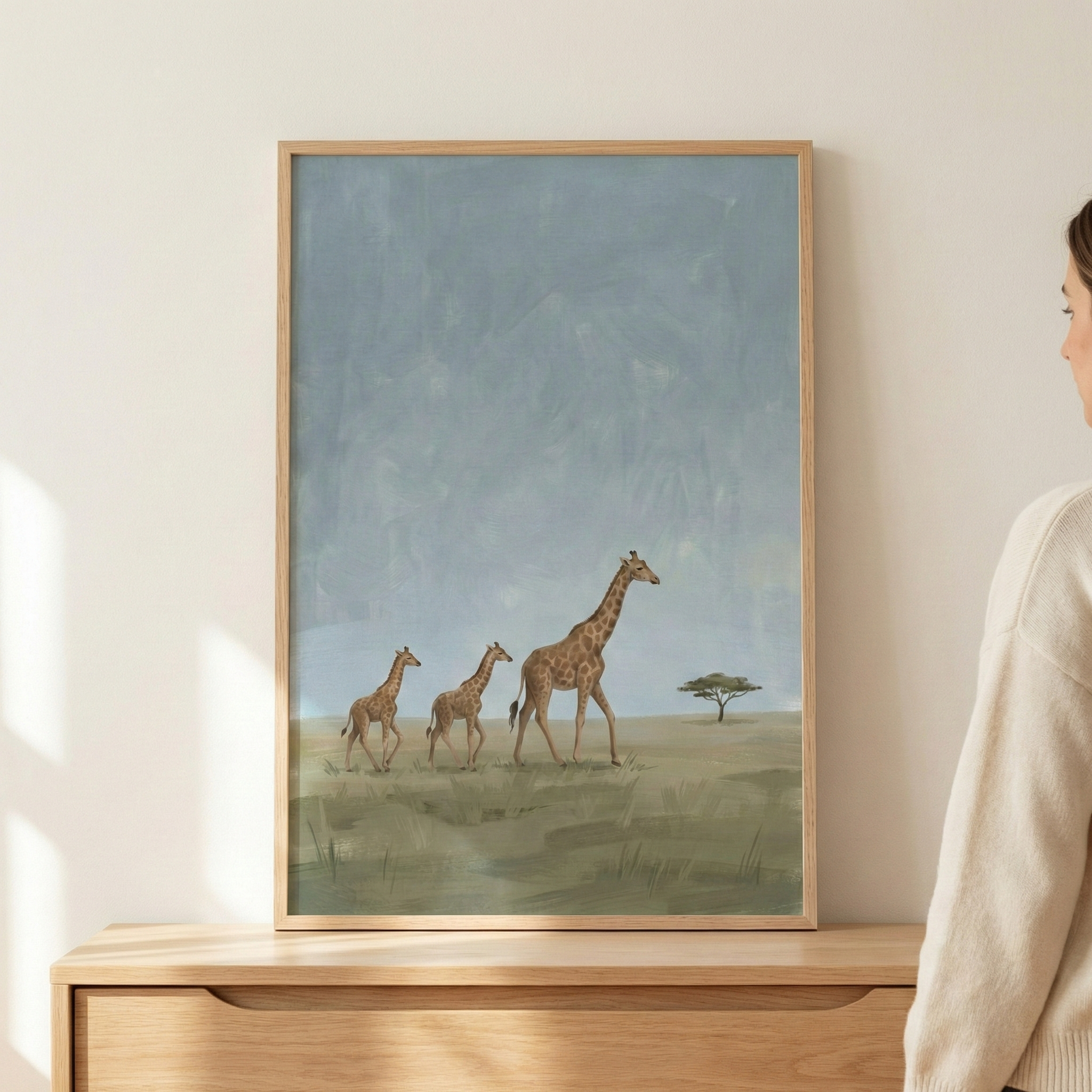 A vertically oriented, light wood-framed art piece hangs on a neutral-toned wall above a light-colored wooden dresser. The artwork within the frame depicts a serene landscape featuring three giraffes—a large adult followed by two smaller ones—walking across a grassy plain. In the distance, a lone, flat-topped tree stands against a vast, atmospheric blue-grey sky. 
