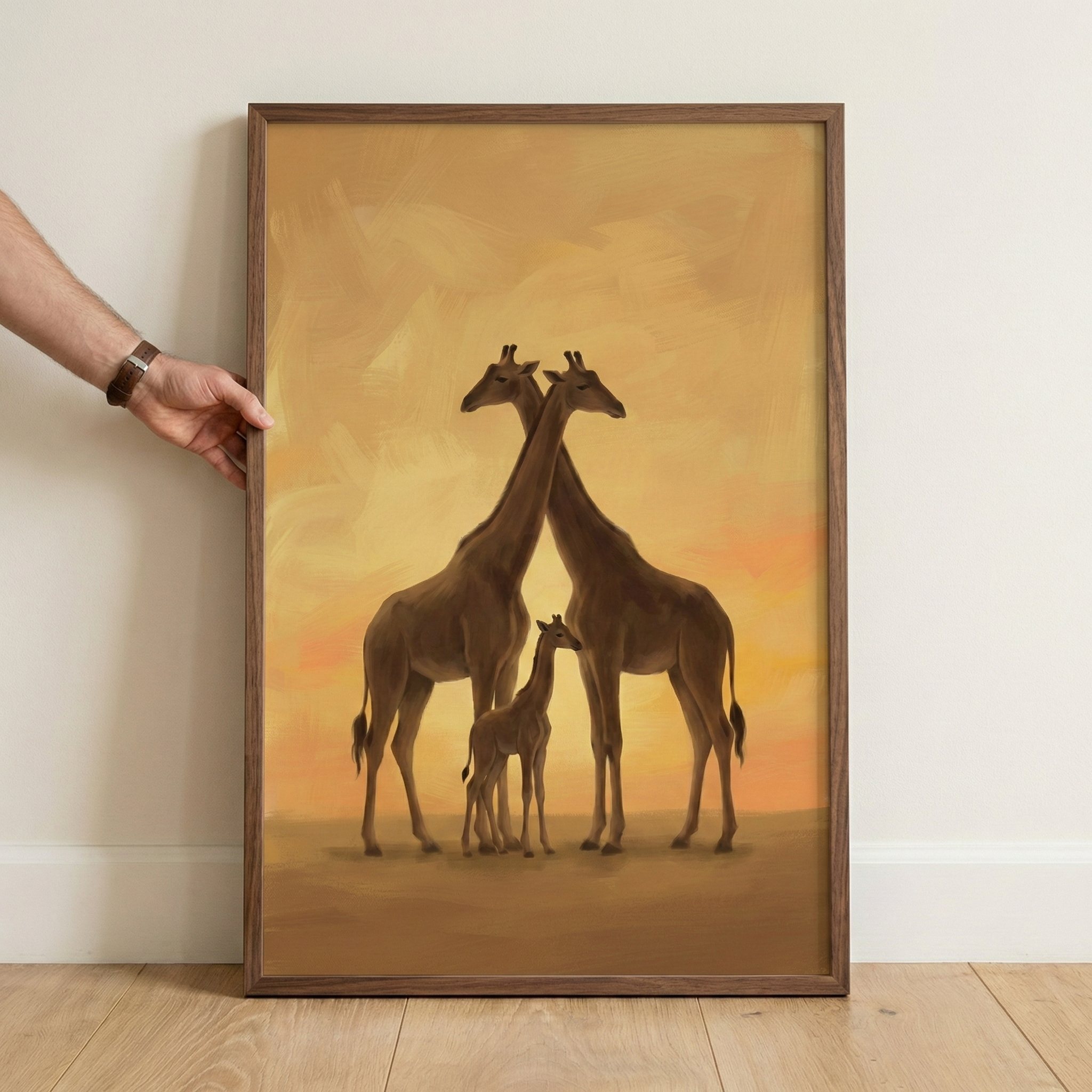 A framed vertical artwork depicts a giraffe family against a warm, golden backdrop. Two adult giraffes stand tall, their necks crossed in a graceful silhouette, while a small youngling stands between them. The scene is rendered in soft, warm tones of amber, yellow, and brown, with visible textured strokes throughout the sky and ground. The art is enclosed in a dark-toned wood frame. A person's hand is visible on the left, holding the frame upright against a plain white wall and light-colored wood flooring.