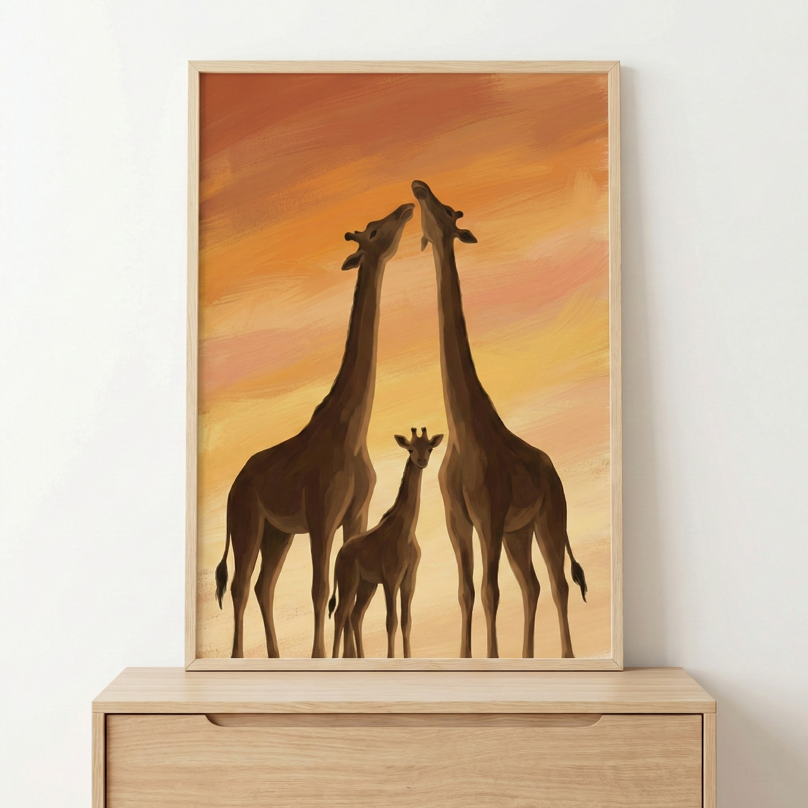A framed piece of art depicts a family of three giraffes set against a warm, textured sunset sky. Two large giraffes stand on either side, their long necks reaching upward and angled toward one another. Between them stands a small giraffe calf, facing the viewer. The sky behind the animals is rendered in horizontal, textured strokes of orange, golden yellow, and soft pink. The artwork is displayed in a light-colored wooden frame, resting on a matching wooden dresser against a plain white wall.