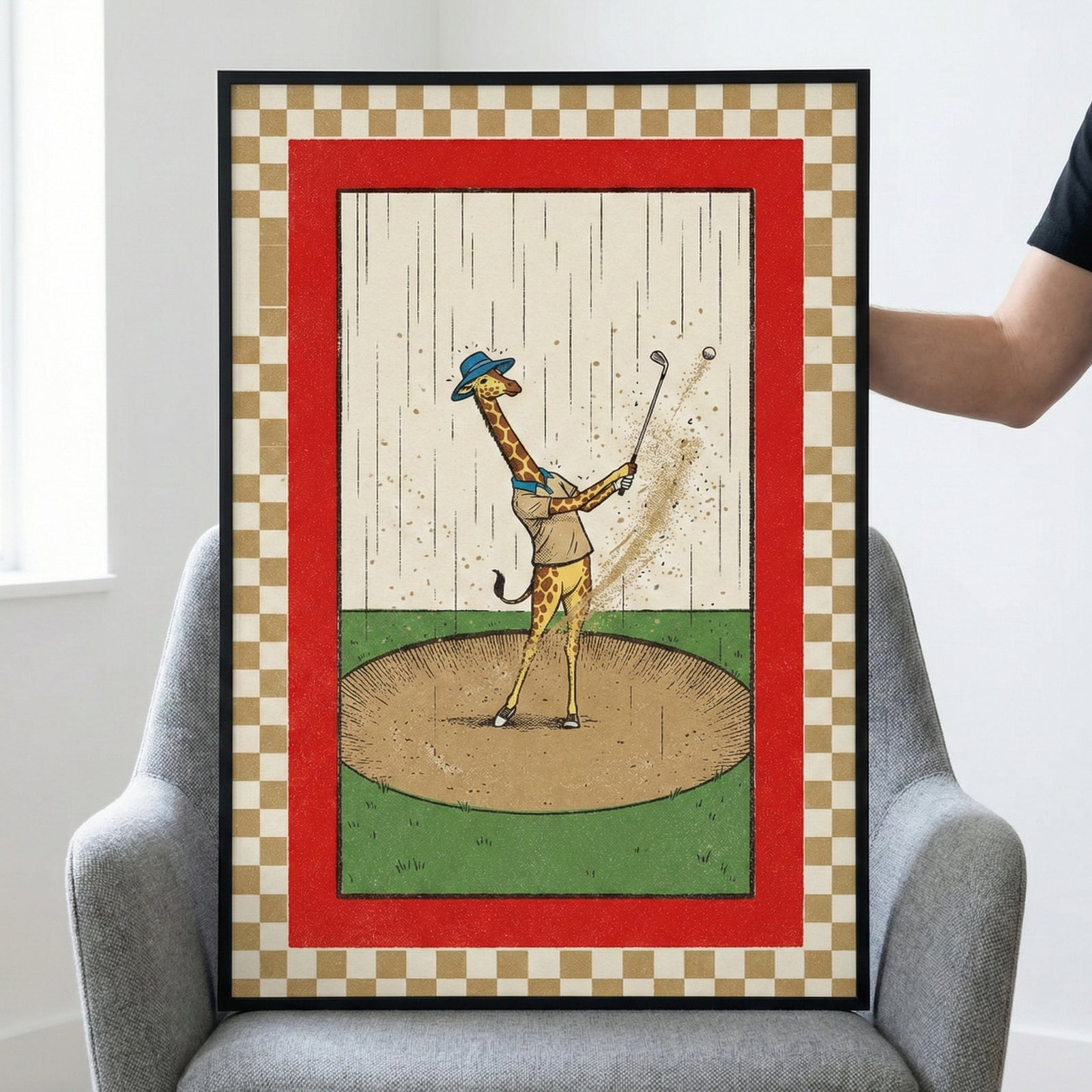 A framed illustration of a giraffe dressed in a blue hat and beige shirt playing golf. The giraffe is mid-swing in a sand trap, with sand and a golf ball flying into the air while rain falls in the background. The central artwork is enclosed by a bold red border and an outer tan and white checkered pattern. The piece is in a black frame and sits on a grey armchair.