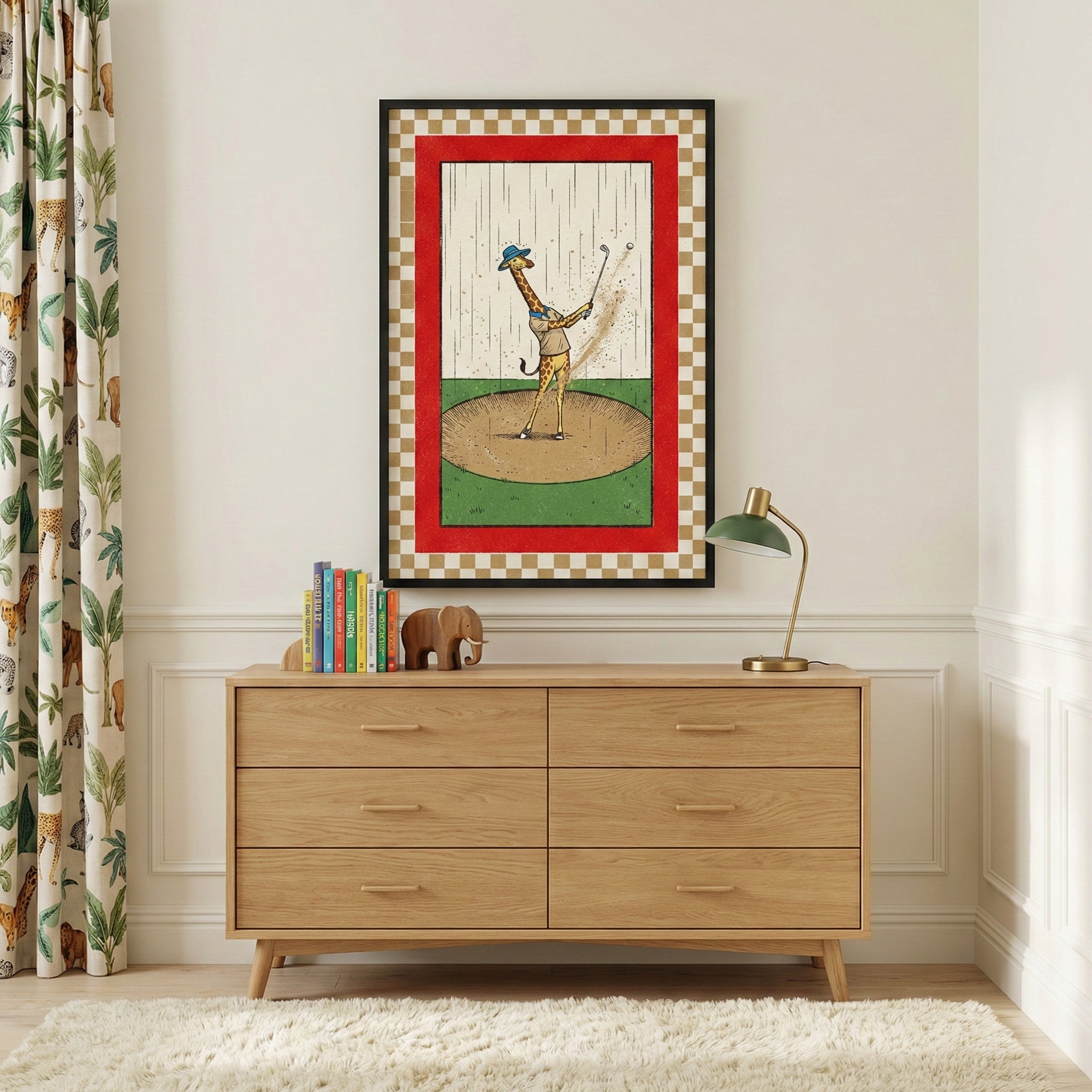 A whimsical child's room features a large framed illustration of a giraffe dressed in a blue hat and vest playing golf in a sand trap under the rain. The artwork is bordered by a bold red frame and a gold and white checkered pattern. It hangs on a white paneled wall above a six-drawer light wood dresser. Atop the dresser sits a collection of colorful books, a small wooden elephant figurine, and a modern gold lamp with a dark green shade. 