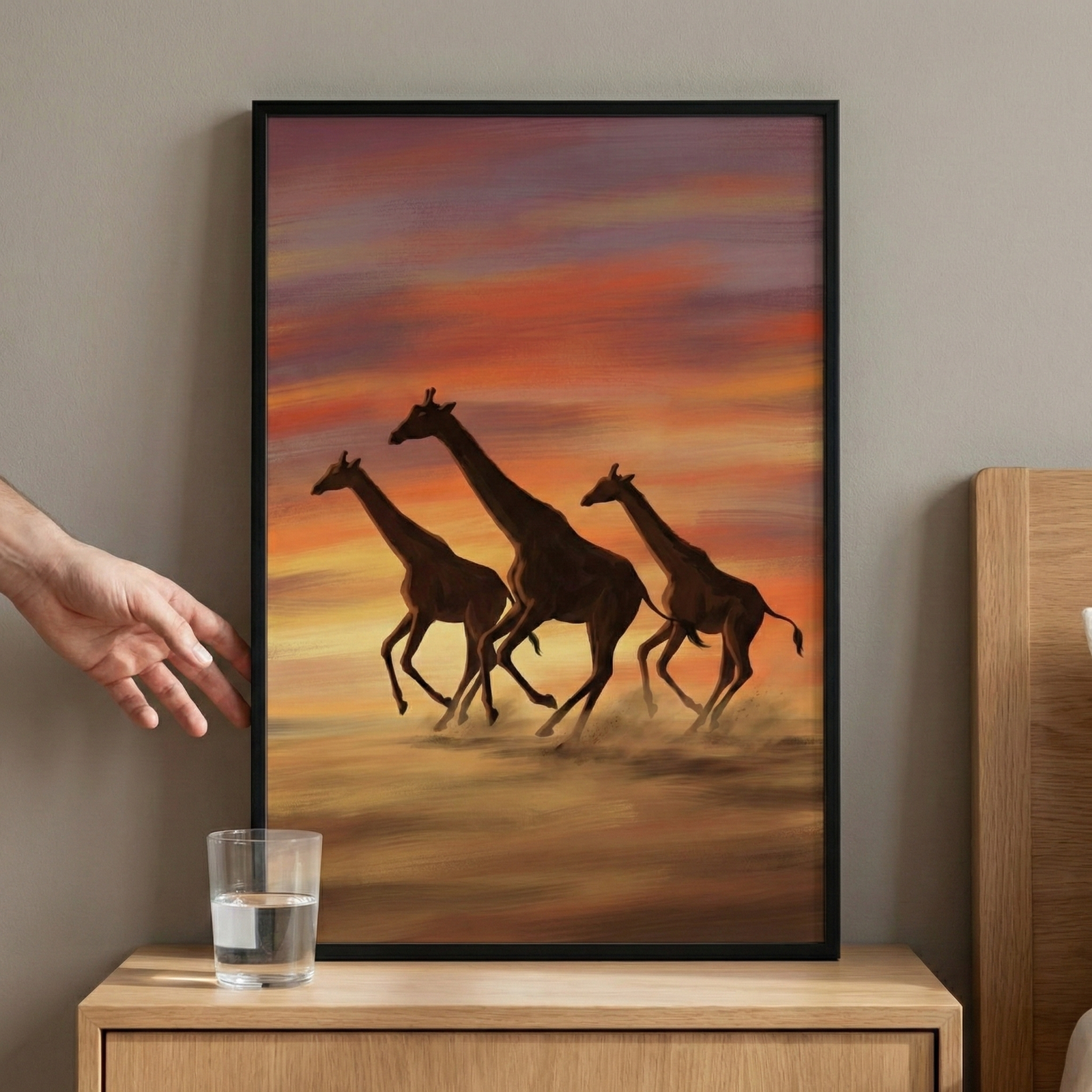 A framed digital artwork captures three giraffes in full gallop across a dusty savanna. The sky is a dramatic palette of orange, red, and purple hues, characteristic of a desert sunset. The scene is full of motion, with the animals depicted in dark tones against the vibrant background, kicking up clouds of dust as they run. The piece is encased in a simple black frame and displayed on a light-colored wall above a wooden bedside table.