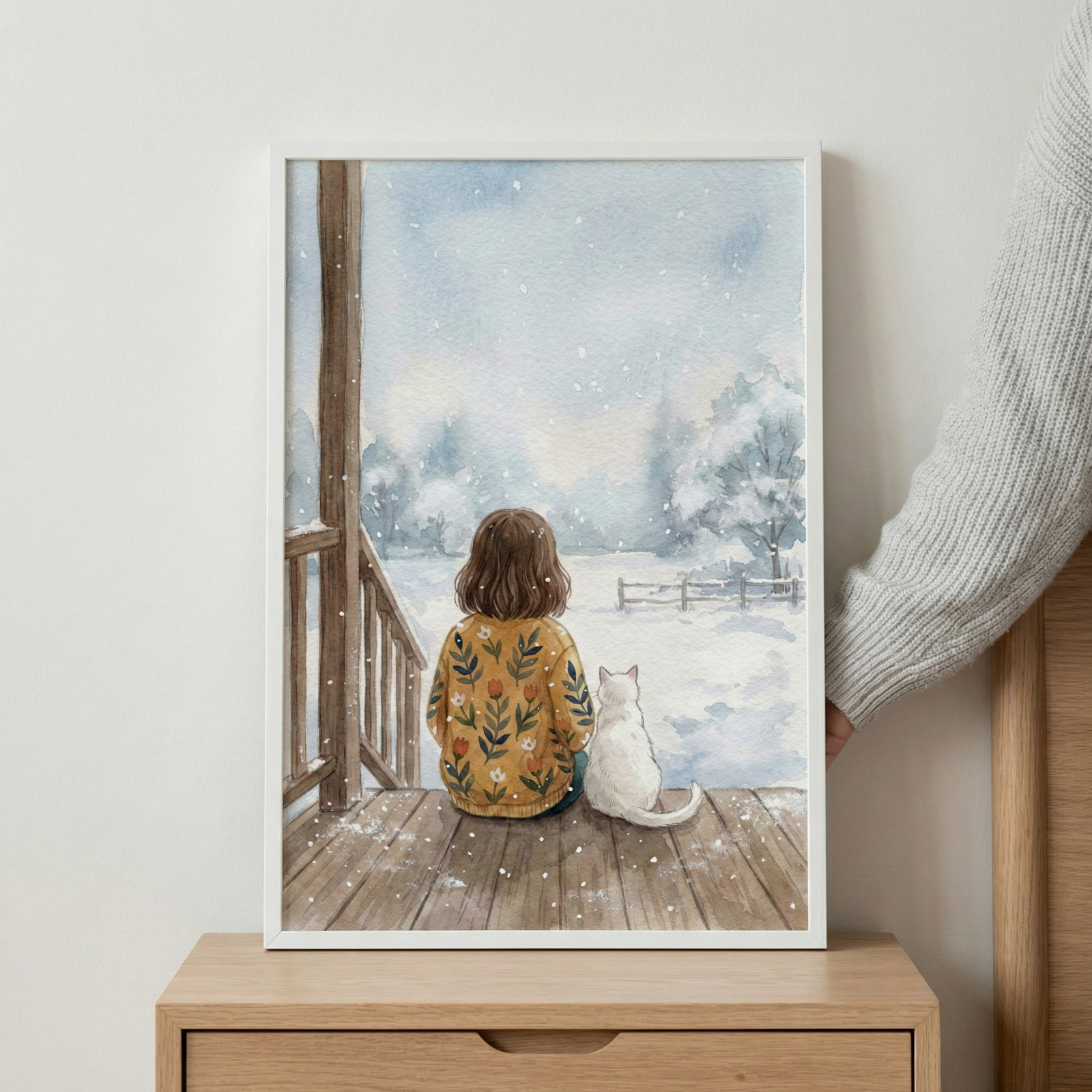 A framed watercolor-style illustration shows a child and a white cat sitting side-by-side on a wooden deck, looking out at a peaceful, snowy field. The child, seen from behind, has brown hair and wears a yellow sweater adorned with colorful flowers. In the distance, a fence and trees are visible through the light snowfall under a soft blue sky. The frame sits on a wooden nightstand against a white wall, with a person's arm in a grey sweater partially visible on the right.