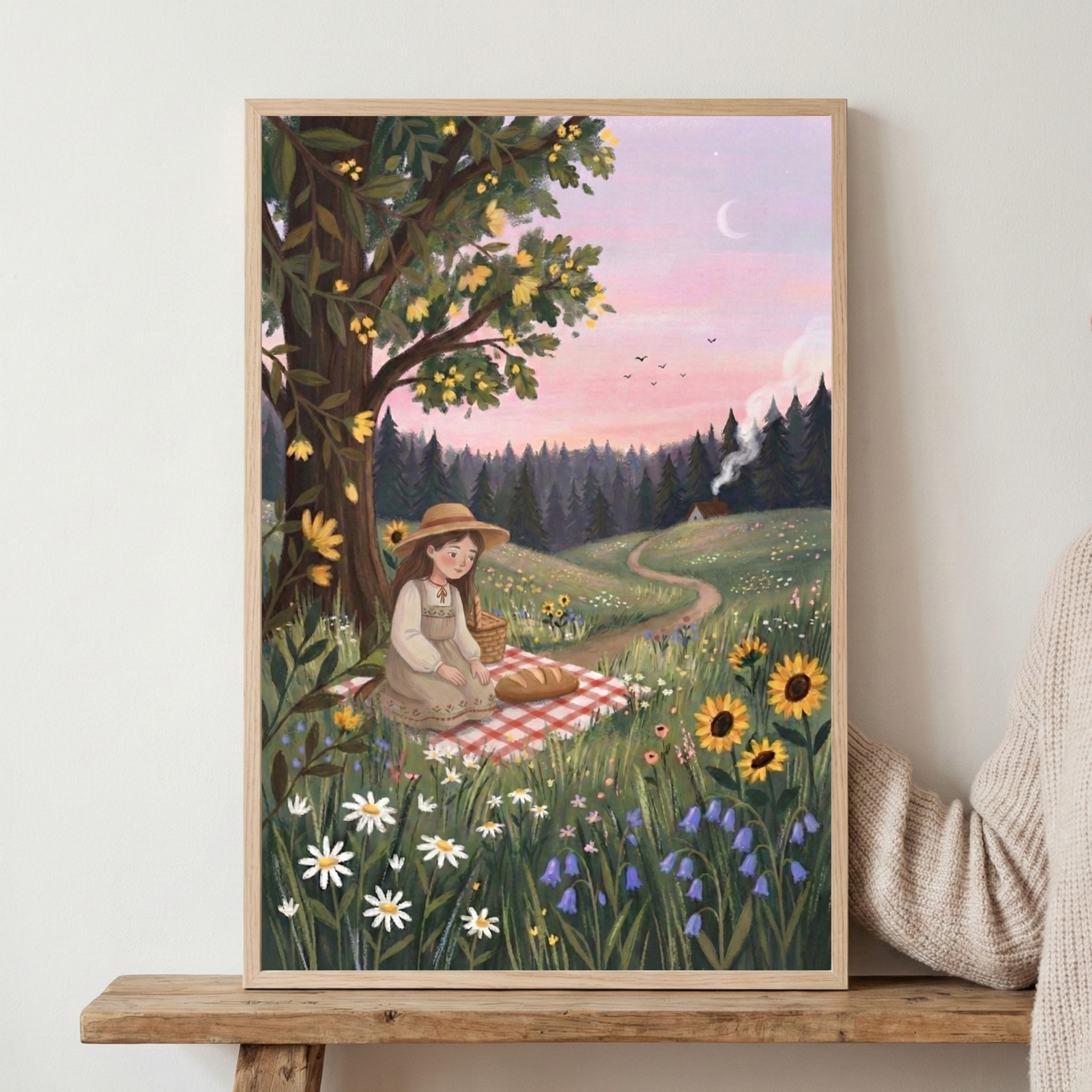 Framed illustration leaning on a wooden surface showing a girl in a straw hat sitting on a red checkered picnic blanket beneath a flowering tree in a wildflower meadow. A winding path leads toward a small cabin in the distance under a pink evening sky with a crescent moon and birds flying overhead.