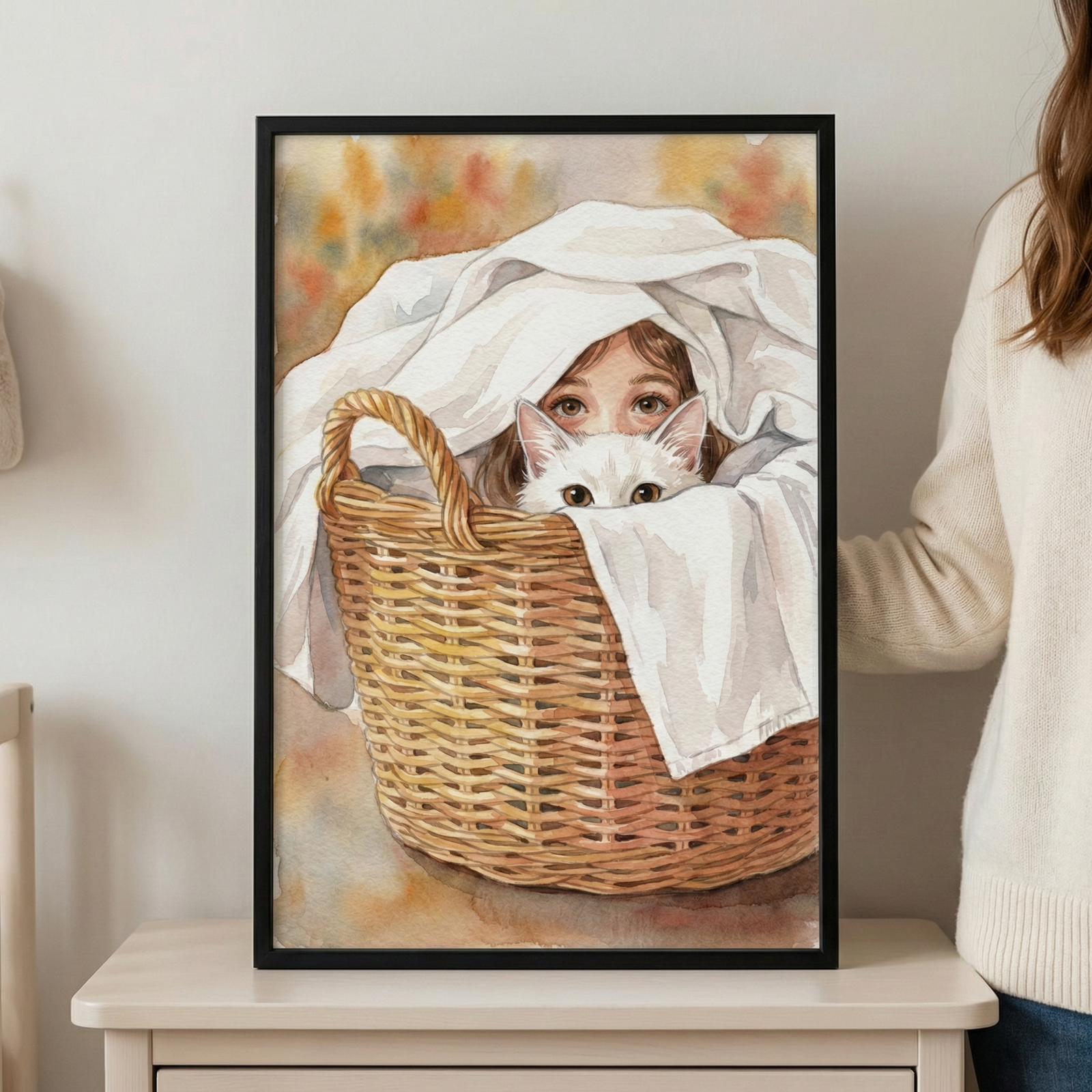 A framed piece of artwork sits on a white surface, held on one side by a person in a cream-colored sweater. The watercolor-style illustration depicts a young girl with large brown eyes peeking out from under a white cloth inside a woven wicker laundry basket. A white cat with dark eyes is also tucked into the basket, peeking out just below the girl. The scene has a soft, whimsical feel with a blurred background of warm, muted colors, all contained within a simple black frame.