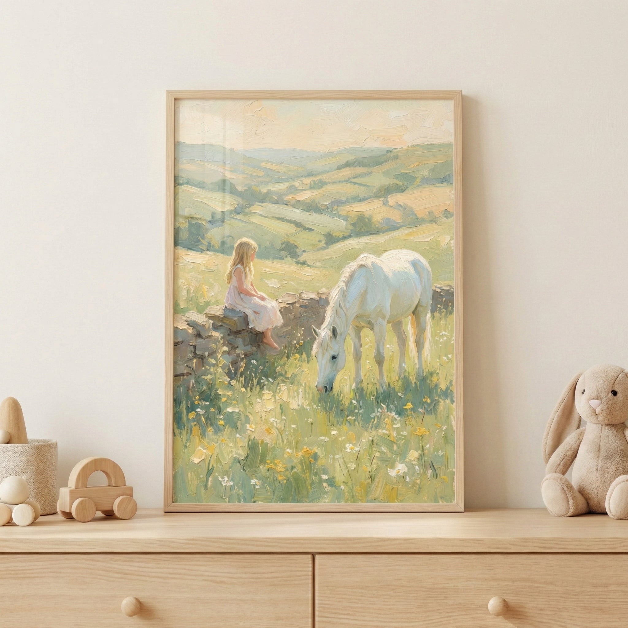 A framed pastel painting of a young girl in a light dress sitting on a stone wall while a white horse grazes in a sunlit wildflower meadow, displayed on a wooden dresser. Soft rolling hills stretch across the background in muted green and golden tones, and simple wooden toys and a plush bunny beside the frame create a calm, storybook inspired nursery setting.