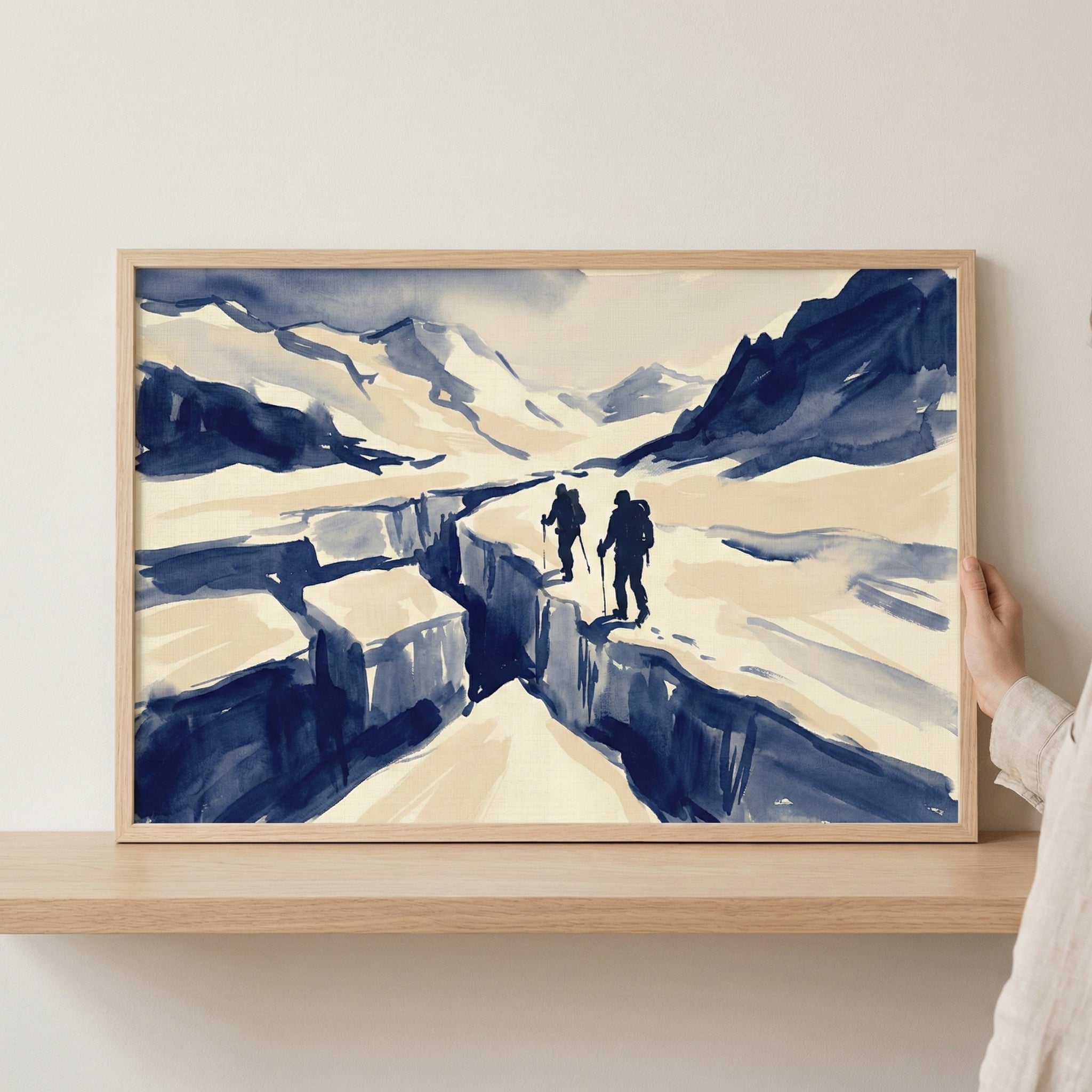 A framed landscape artwork in a minimalist watercolor style, featuring two silhouetted figures with backpacks and walking sticks trekking across a snowy mountain range. The hikers are positioned on the edge of a deep, navy blue crevasse. The scene is created with bold, expressive strokes in shades of blue and cream. The light wood frame rests on a matching wooden shelf, with a hand visible on the right side holding the frame against a neutral-toned wall.
