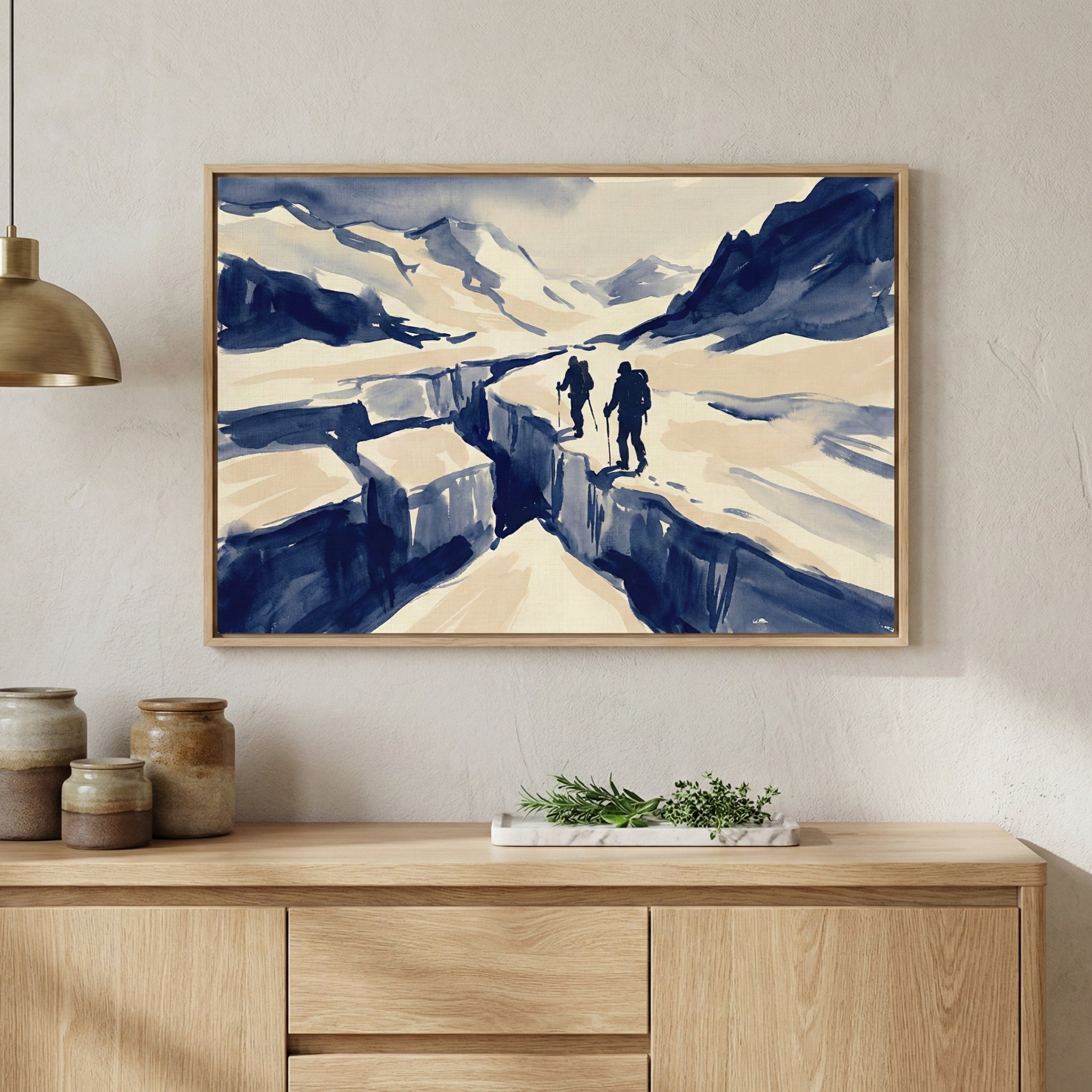 A framed piece of minimalist artwork in a fluid, watercolor style hangs on a textured neutral wall. The art depicts two silhouetted hikers with backpacks and trekking poles walking along the edge of a deep, dark blue crevasse in a vast, snowy mountain landscape. The color palette is limited to shades of navy blue and cream. The light wood frame is mounted above a light-colored wooden console table, which holds three earthy ceramic jars and a small marble tray with green sprigs. A portion of a gold pendant l