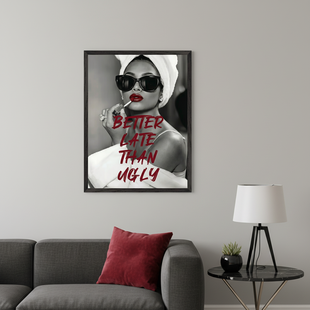 Modern living room with a dark gray sectional sofa. Framed black and white wall art hangs above the sofa, featuring a woman in sunglasses applying red lipstick, with red text "BETTER LATE THAN UGLY" overlaid. A black marble side table with a lamp is on the right.
