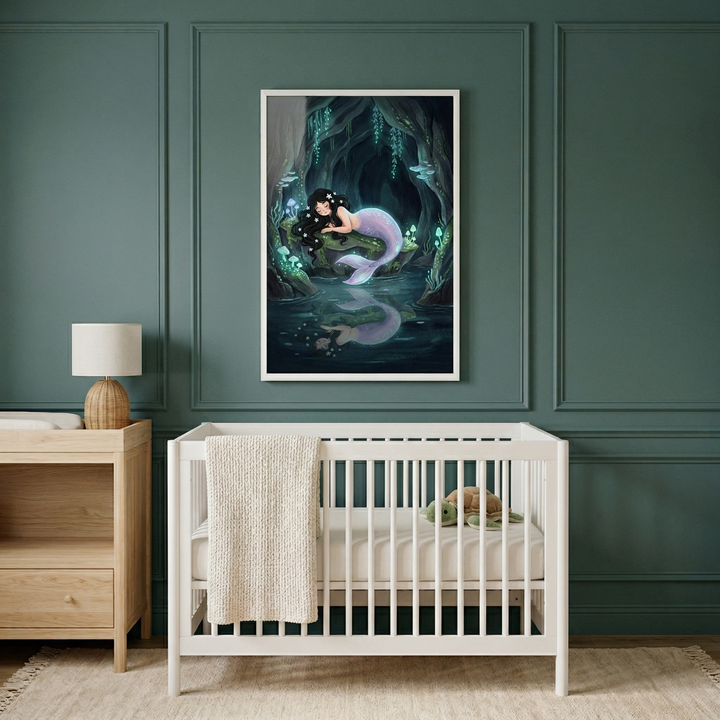 A modern nursery features dark teal paneled walls and a white wooden crib centered in the frame. The crib contains a light mattress and a plush turtle toy, with a textured beige blanket draped over the side. To the left is a light wood dresser topped with a small lamp. Hanging above the crib is a large framed illustration of a dark-haired mermaid with a purple tail in a glowing, magical cave, resting near water that reflects her image.