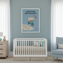 Modern nursery with a white crib against a blue wall. A framed print hangs centered above the crib, featuring text "EXECUTING SKULDUGGERY." and a cartoon ibex riding a blue BMX bike down stairs. A wooden dresser is to the left and a light blue armchair is to the right.