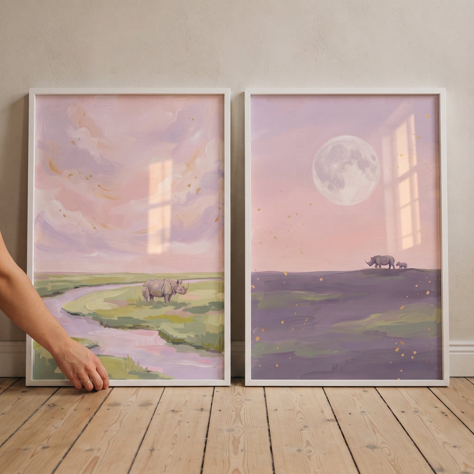 Gold flecked pink rhino wall art set of 2 features rhinos in soft pastel landscapes under a pink sky with a river and moon.
