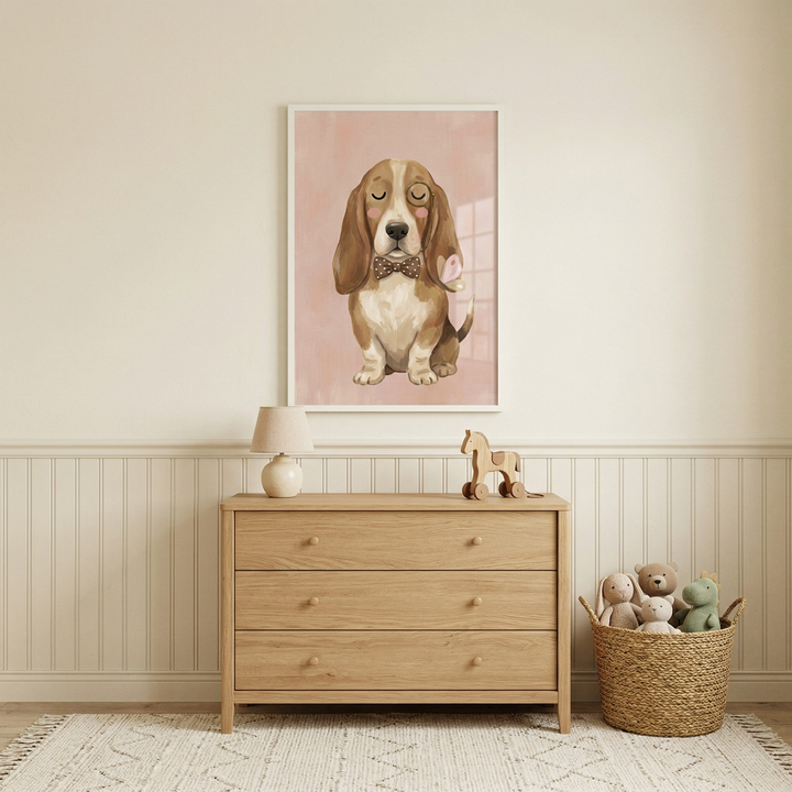 A light and airy nursery features a piece of framed art hanging on a beige wall above a wooden dresser. The artwork depicts a cute, brown and white dog with long ears, sitting and wearing a polka-dot bowtie with its eyes closed. Below the art, a light wood dresser holds a small table lamp and a wooden horse toy. To the right of the dresser, a woven basket is filled with plush animal toys. The room has light-colored wooden paneling on the lower half of the walls and a patterned rug on the floor.