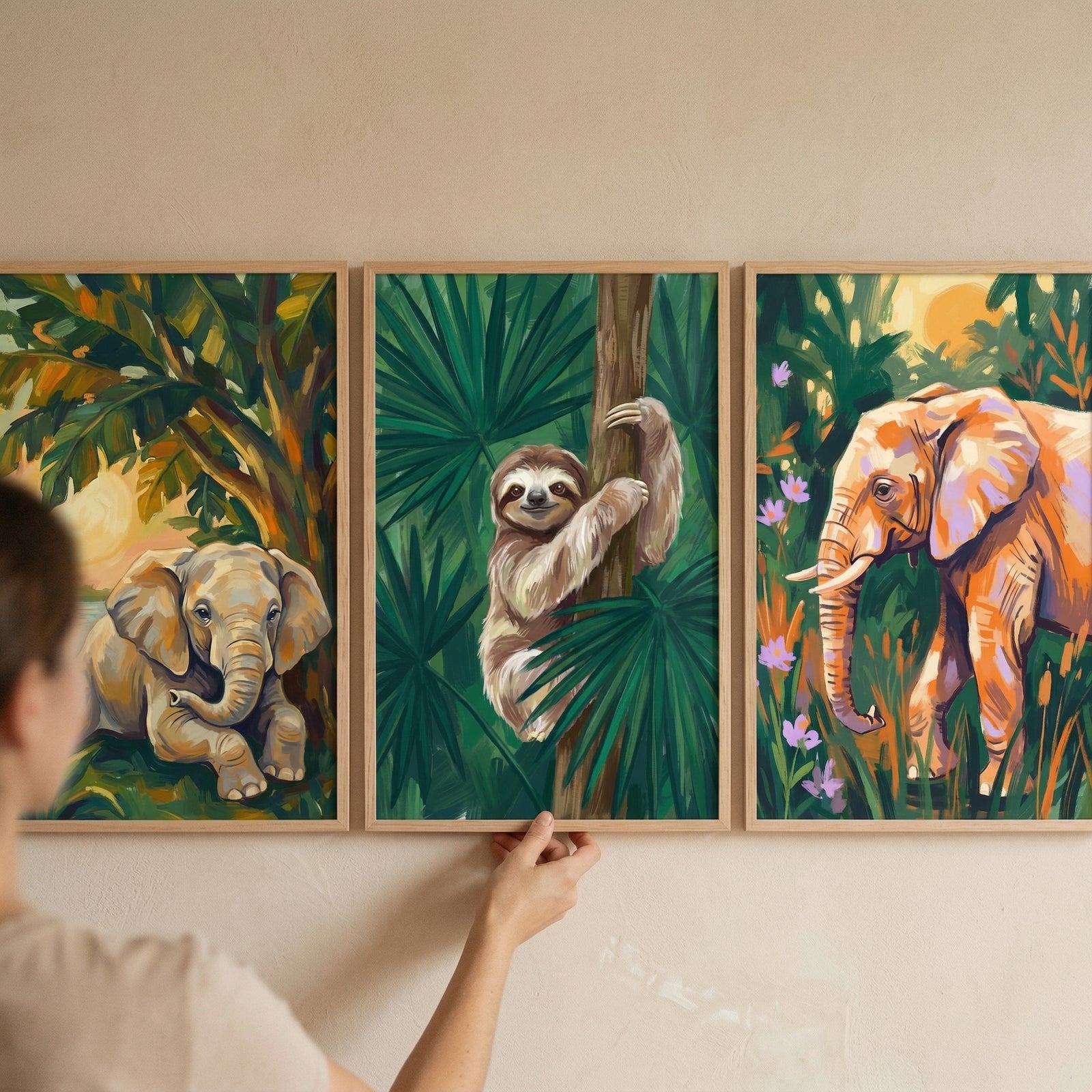 Three framed jungle animal paintings hang on a wall while a person adjusts the center frame. The artwork shows a baby elephant resting under trees, a smiling sloth clinging to a tree trunk surrounded by green leaves, and a standing elephant in a colorful tropical setting.