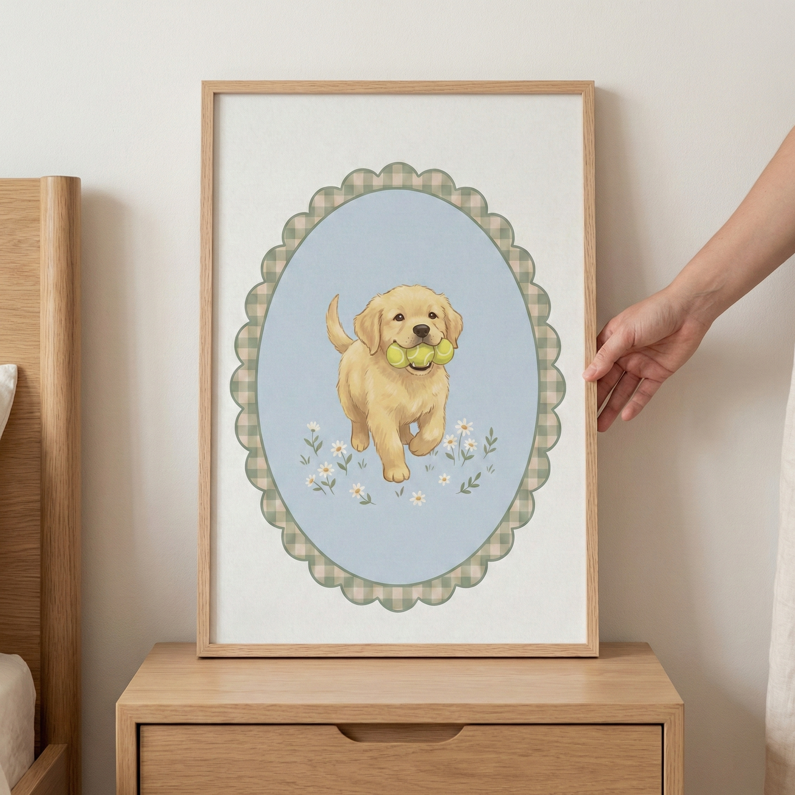 A rectangular wooden frame rests on a light-colored wooden bedside table, held by a hand on the right. Inside the frame is an illustration of a fluffy, golden-colored puppy. The puppy is featured within a scalloped, oval-shaped border with a green and white checkered pattern. Set against a soft blue background, the puppy is walking forward while carrying three green tennis balls in its mouth. Small white daisies are scattered at its feet. The scene is set against a plain, neutral wall.