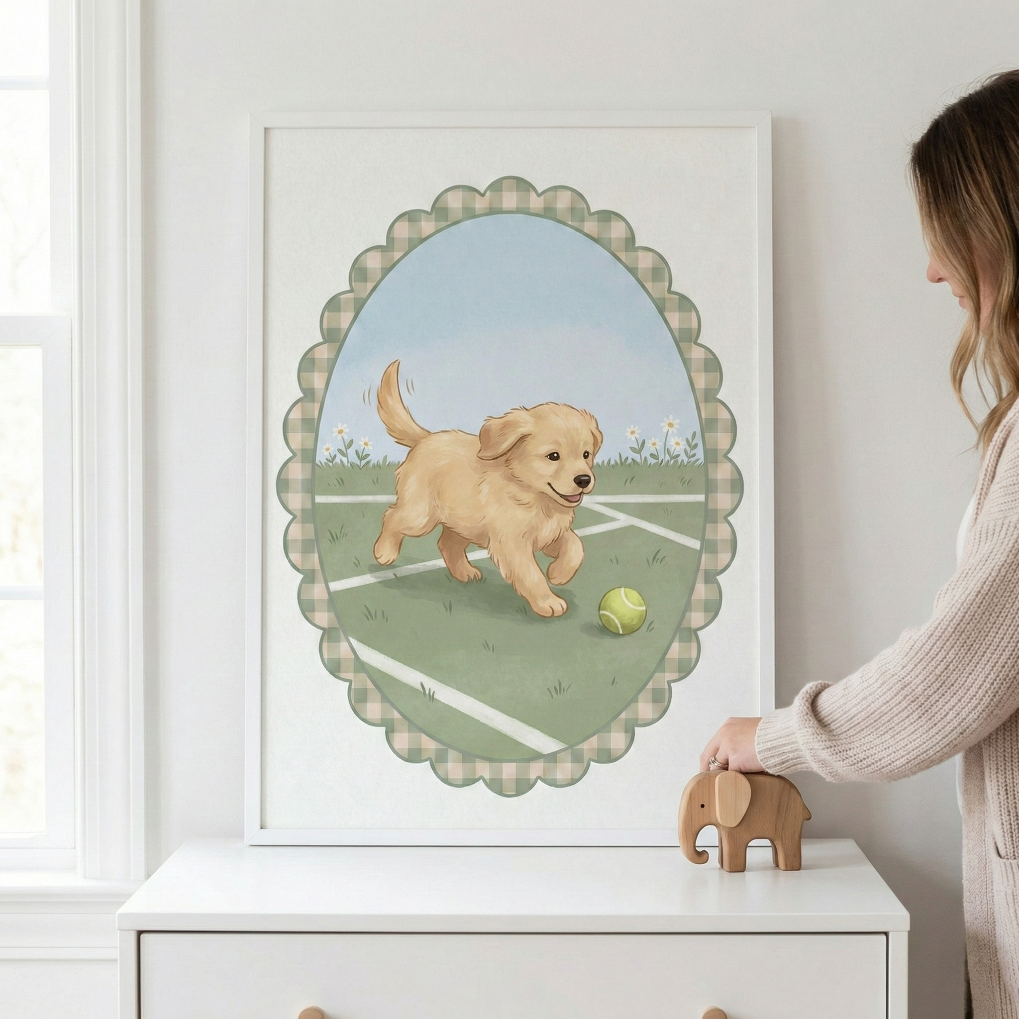 A framed illustration depicts a golden retriever puppy running towards a tennis ball on a grassy court with white daisies. The art is centered in a scalloped oval frame with a green gingham pattern. The frame hangs on a white wall above a white dresser. A person's hand is shown touching a small wooden elephant figurine resting on the dresser. A sunlit window is partially visible on the left.