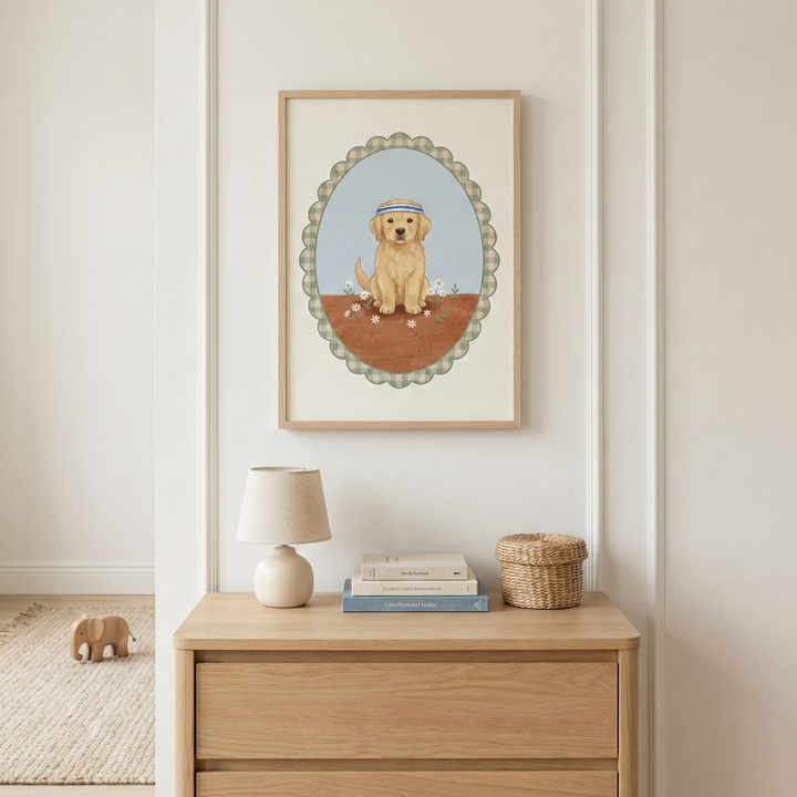 A framed illustration hangs on a white wall above a light wood dresser. The artwork features a golden-colored puppy wearing a blue and white striped headband, sitting in a field of small white flowers. The puppy is centered within an oval frame with a green gingham border. On the dresser sits a small beige lamp, a stack of books, and a round woven basket. A wooden elephant toy is visible on a neutral-toned rug in the lower-left corner.