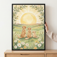 A serene watercolor illustration depicts three golden-furred dogs of varying sizes—an adult, a younger dog, and a puppy—sitting together on a grassy hill, viewed from behind. They gaze toward a large, soft yellow sun rising over rolling green hills. The foreground is dotted with white and yellow wildflowers. Two arching leafy branches frame the scene at the top. The artwork is shown in a black frame, held against a white wall and resting on a wooden surface.