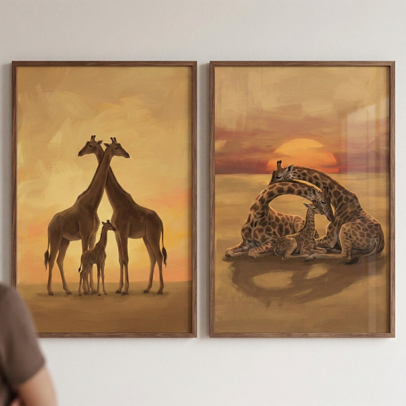 Two framed giraffe paintings show calm family scenes in warm sunset colors. One artwork shows two tall giraffes standing close together with a small calf between them under a golden sky. The other shows two adult giraffes curving their long necks protectively around a resting calf with the sun setting behind them.