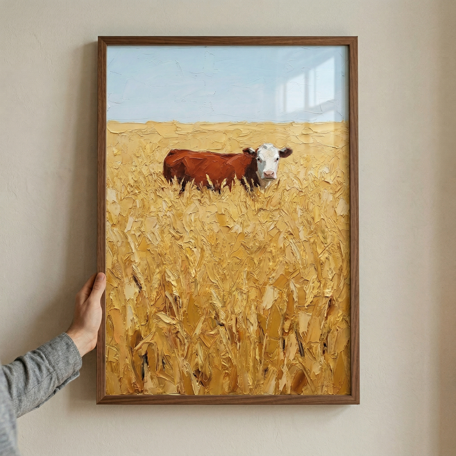 A vertical textured artwork is displayed within a dark wooden frame, held against a neutral beige wall by a person's hand on the left. The scene within the frame captures a brown and white cow standing in a vast field of golden wheat. The sky above is a pale, muted blue with broad, horizontal strokes of white.
The entire surface of the artwork is characterized by a heavy, impasto-like texture, with thick ridges and clumps of pigment that give the scene a three-dimensional quality. 