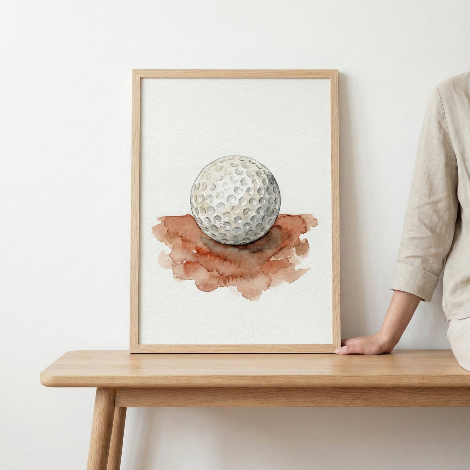 Minimalist watercolor painting of a single golf ball resting on a splash of brown earth tones. The soft brushwork highlights the texture of the dimples, symbolizing simplicity in the sport of golf.