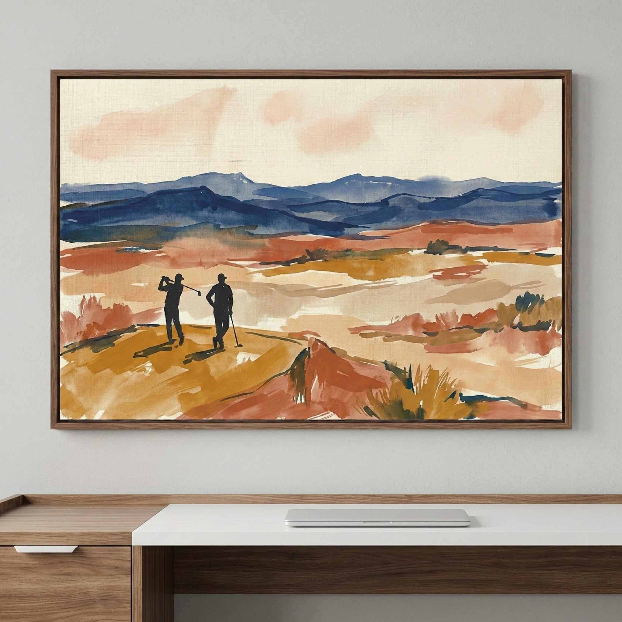 A piece of framed wall art depicting two silhouetted figures on a golf course set against a landscape of rolling blue mountains. The artwork is created with expressive, broad brushstrokes in a palette of terracotta, ochre, navy, and soft pink. One figure is captured in the middle of a swing, while the other stands beside them. The sky is a pale cream color with soft, wispy clouds. The frame is a thin dark wood, and the piece hangs above a modern white desk with a wooden drawer and a closed laptop.