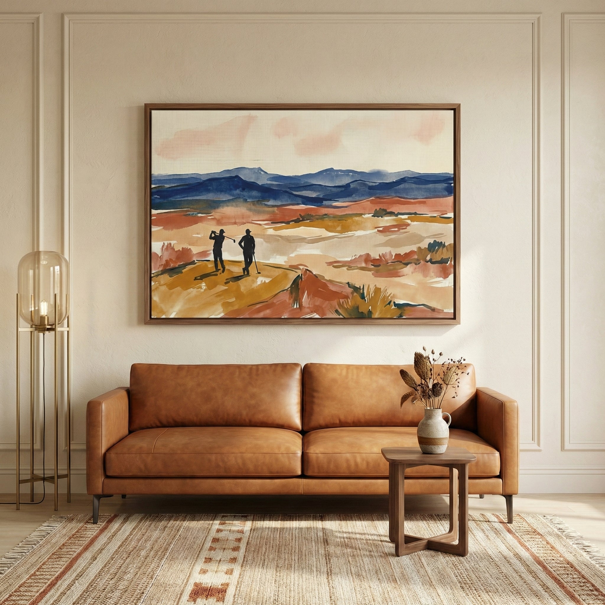 A modern, warm-toned living room features a large framed artwork centered on a wall with decorative molding. The artwork depicts two silhouetted figures standing on a golden-brown terrain with a backdrop of layered blue mountains and a soft pink sky. Below the art is a cognac leather two-seater sofa. To the left is a tall, slender metal floor lamp with a cylindrical glass shade. To the right, a small dark wood side table holds a ceramic vase with dried botanical elements. The room is completed by a patterne