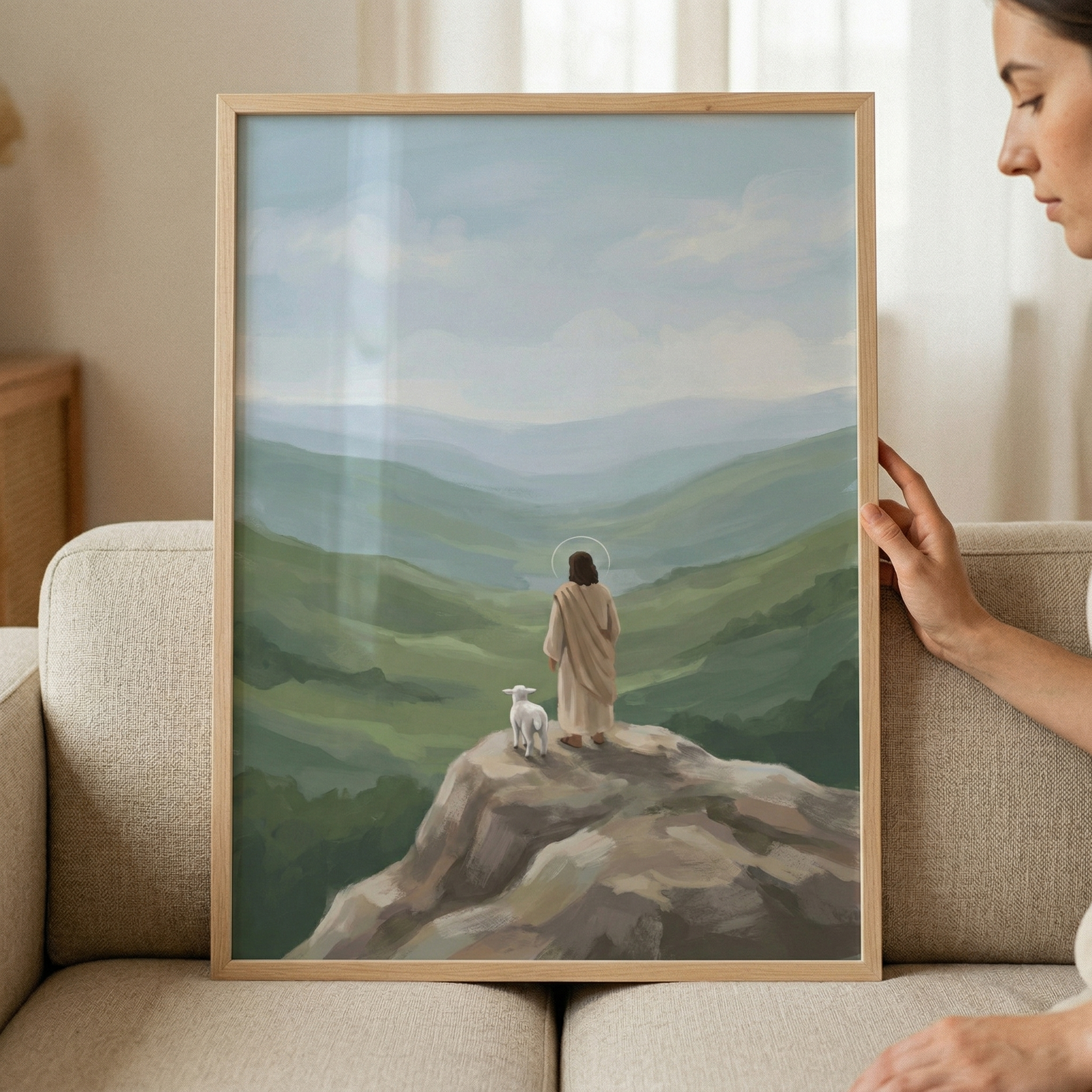 A light wood-framed work of art held by a person on a beige sofa. The illustration depicts Jesus from behind, standing on a rocky outcrop. He wears a light tan robe and has a simple halo. A small white lamb stands beside him. They are looking out over an expansive valley filled with layered green hills under a soft blue sky with light clouds. The art has a soft, stylized aesthetic with subtle textures.