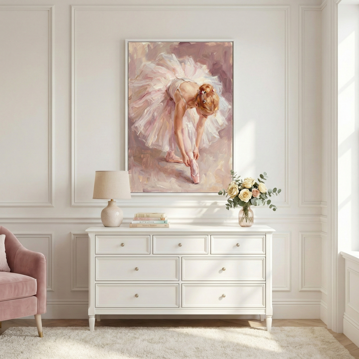 A bright, elegant room features white paneled walls and a large, framed canvas. The artwork depicts a ballerina in a voluminous pink tutu, bending over to fasten her pointe shoes in a soft, impressionistic style. Below, a classic white dresser with seven drawers holds a cream-colored lamp, a stack of books, and a vase of pale roses. To the left is the edge of a plush pink armchair. A soft, light-colored rug covers the floor, illuminated by natural light from a window.